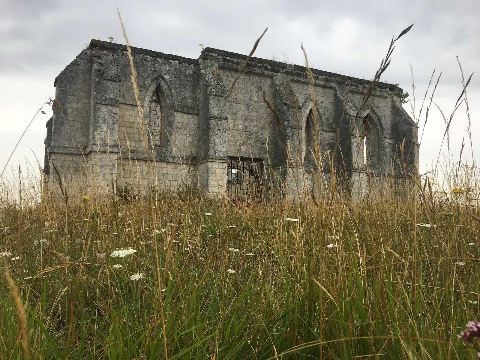 The roofless stone shell of a ruined chapel, its pointed Gothic window openings empty of glass, rises above a meadow of tall grass and wildflowers.