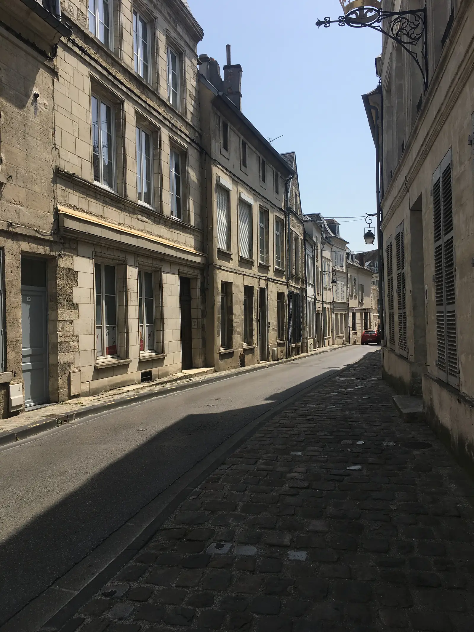 A narrow street of tall stone townhouses with shuttered windows curves gently uphill, a red vehicle parked in the distance.