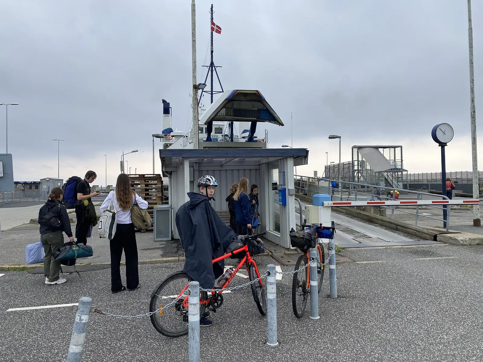 Foot passengers and two loaded bikes waiting at a ferry ramp beside a kiosk and barrier, the ferry's bridge and Danish flag behind.