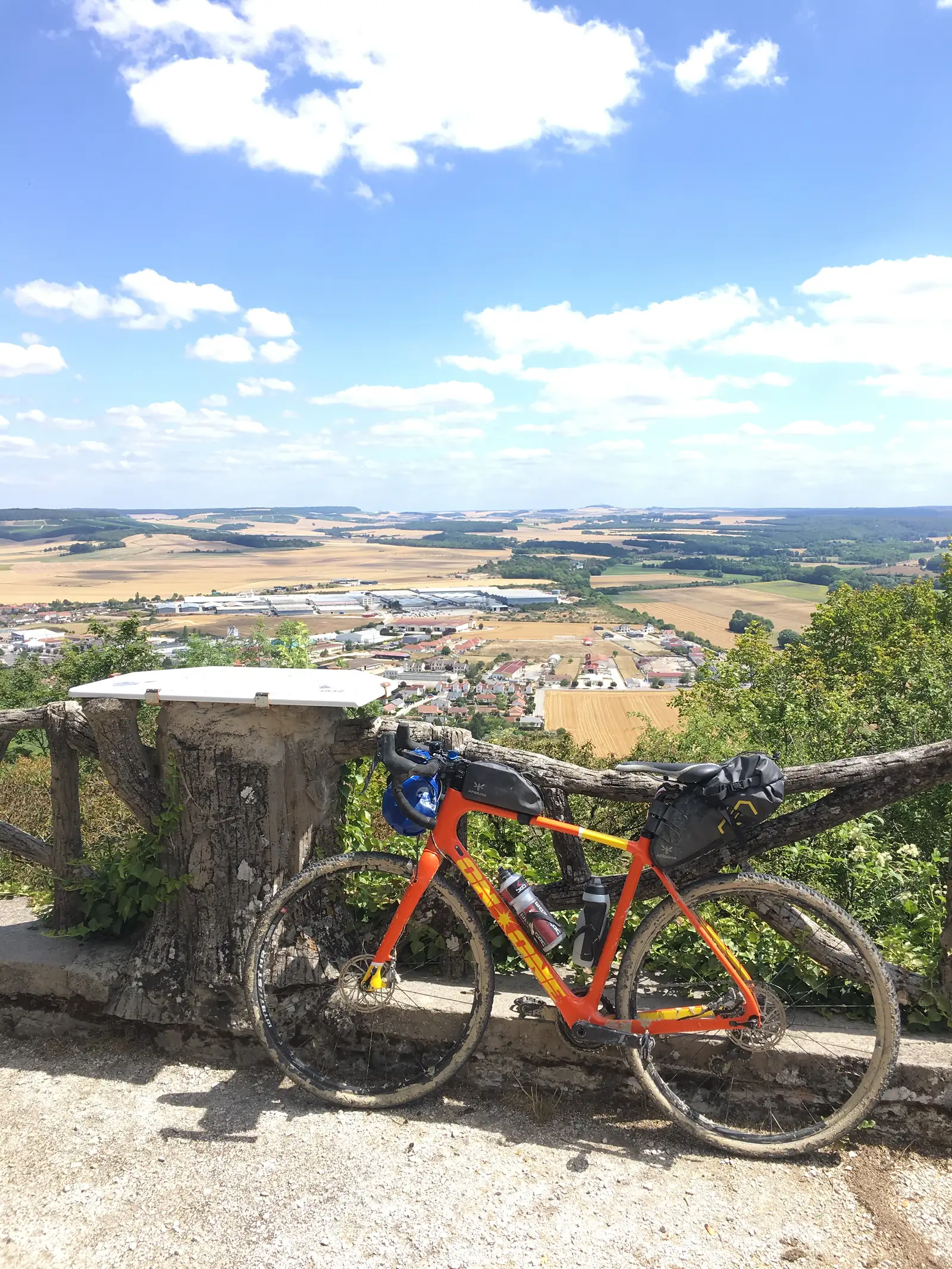 A mud-spattered gravel bike, laden with bikepacking bags and water bottles, stands at a viewpoint overlooking a town in the valley below.