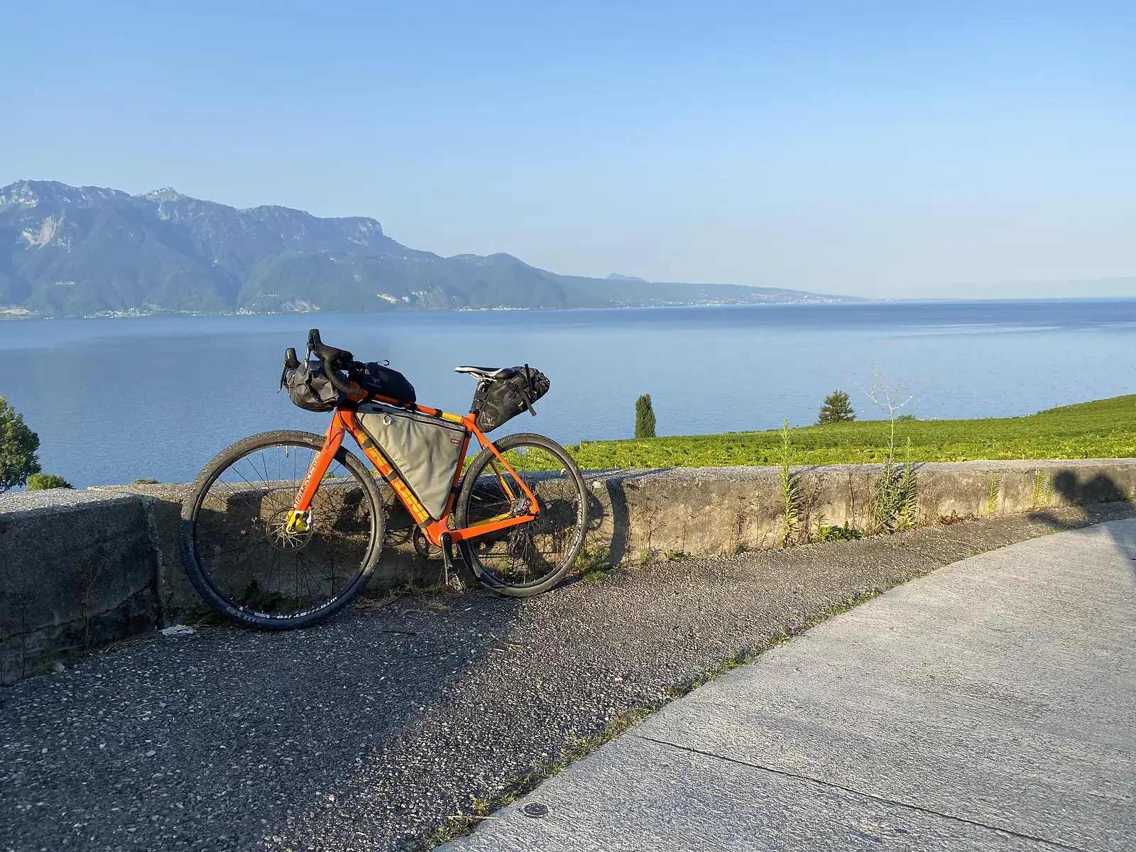 A loaded orange gravel bike with black frame and handlebar bags leans against a low stone wall above Lake Geneva, vineyards and mountains stretching into the haze beyond.