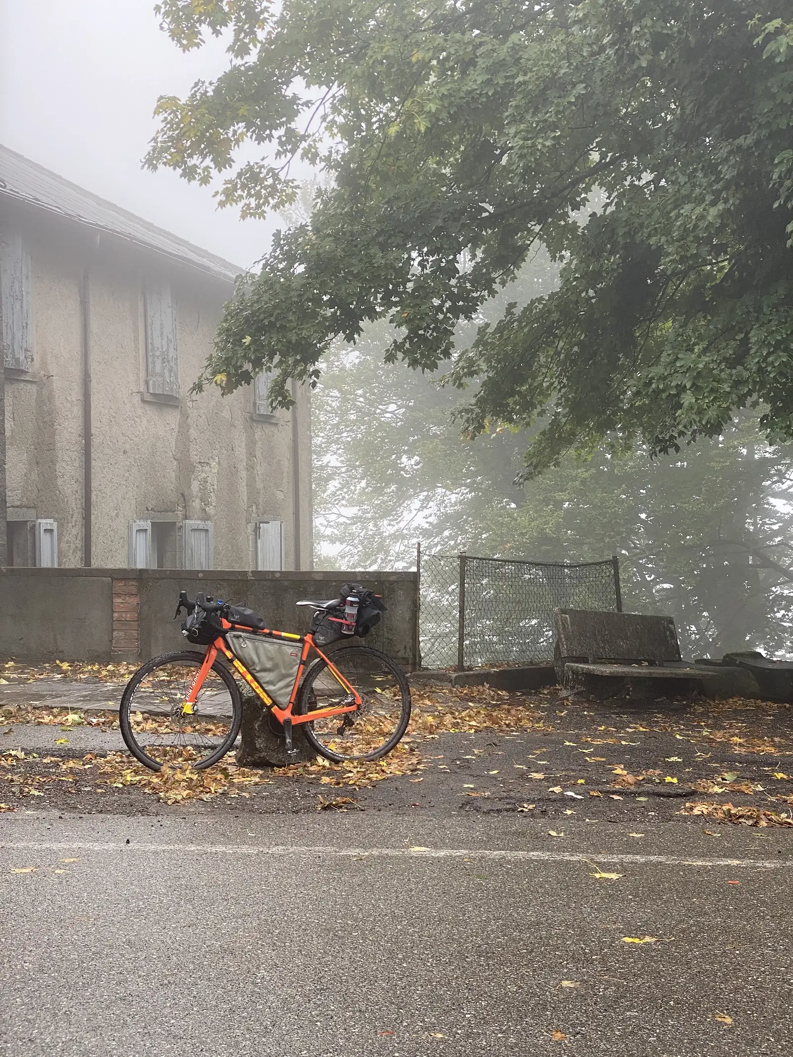 A loaded orange bikepacking bike leans against a stone building on a leaf-strewn forecourt, mist hanging heavily among the trees behind.