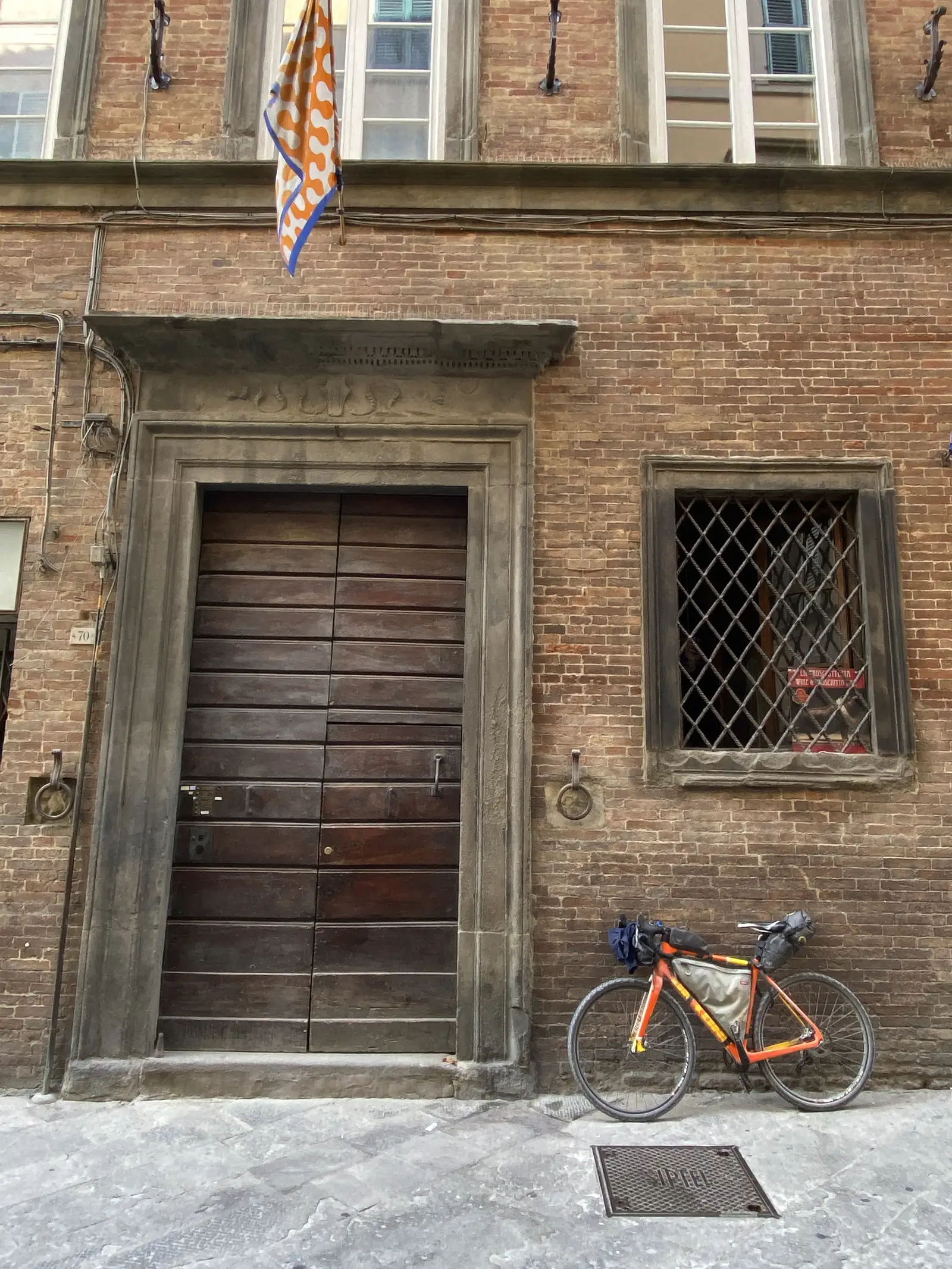 An orange bikepacking bike leaning against a brick townhouse wall beside a grated window and a large wooden double door numbered 70.