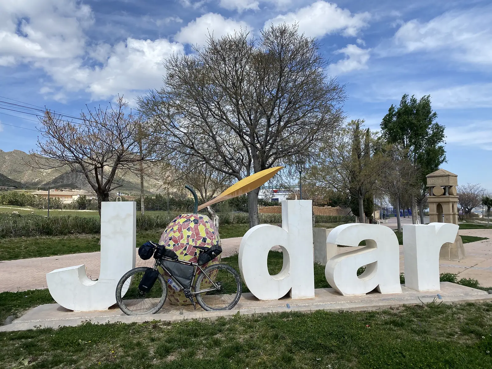 A loaded bikepacking bike leaning against giant letters spelling "Jodar" in a roadside park, with a mosaic olive sculpture and mountains beyond.