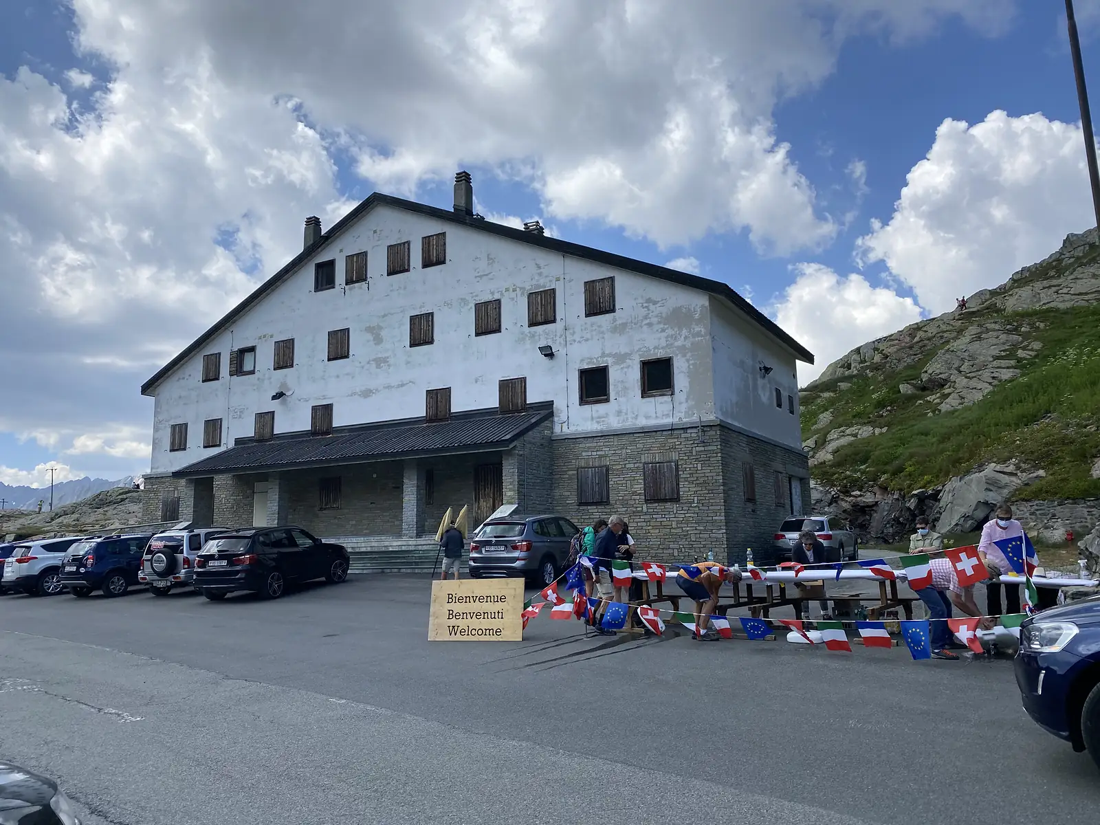 
A weathered alpine building with a wooden "Bienvenue Benvenuti Welcome" sign out front, where people decorate picnic tables with Swiss, Italian, French and EU flags.
