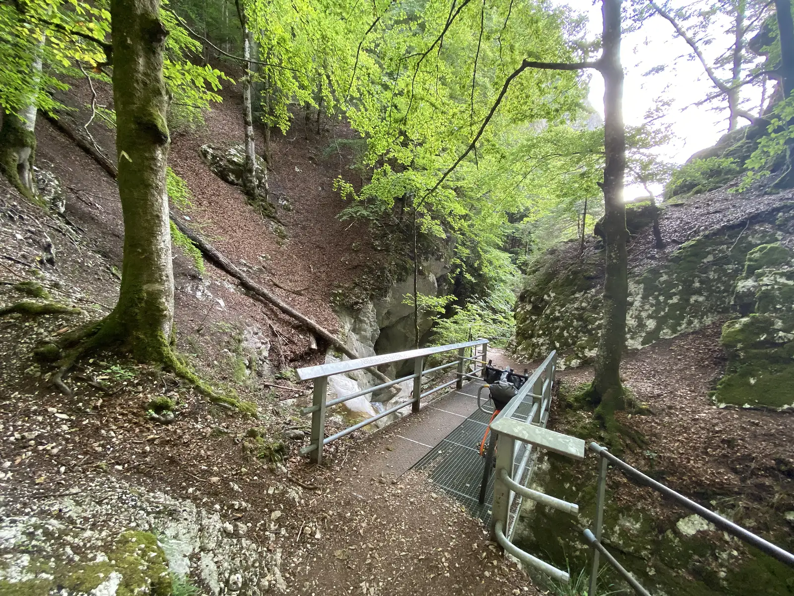 A metal-grate footbridge crossing a narrow limestone gorge, with a loaded bike's handlebar bag visible against the railing and a rock archway ahead.