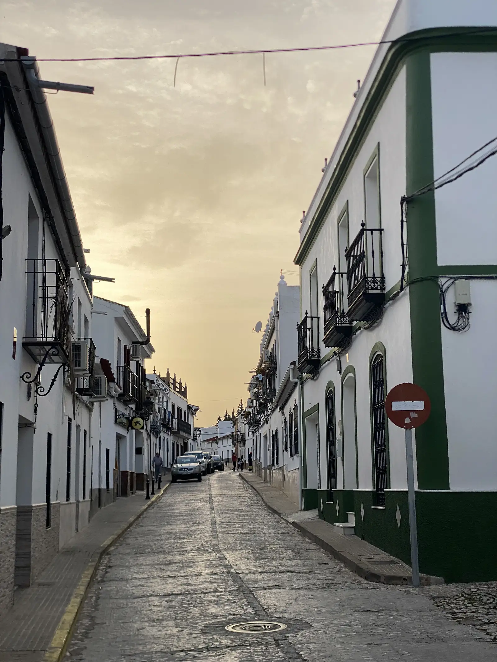 A cobbled street of whitewashed houses with black iron balconies running towards a golden evening sky, a green-painted corner building on the right.