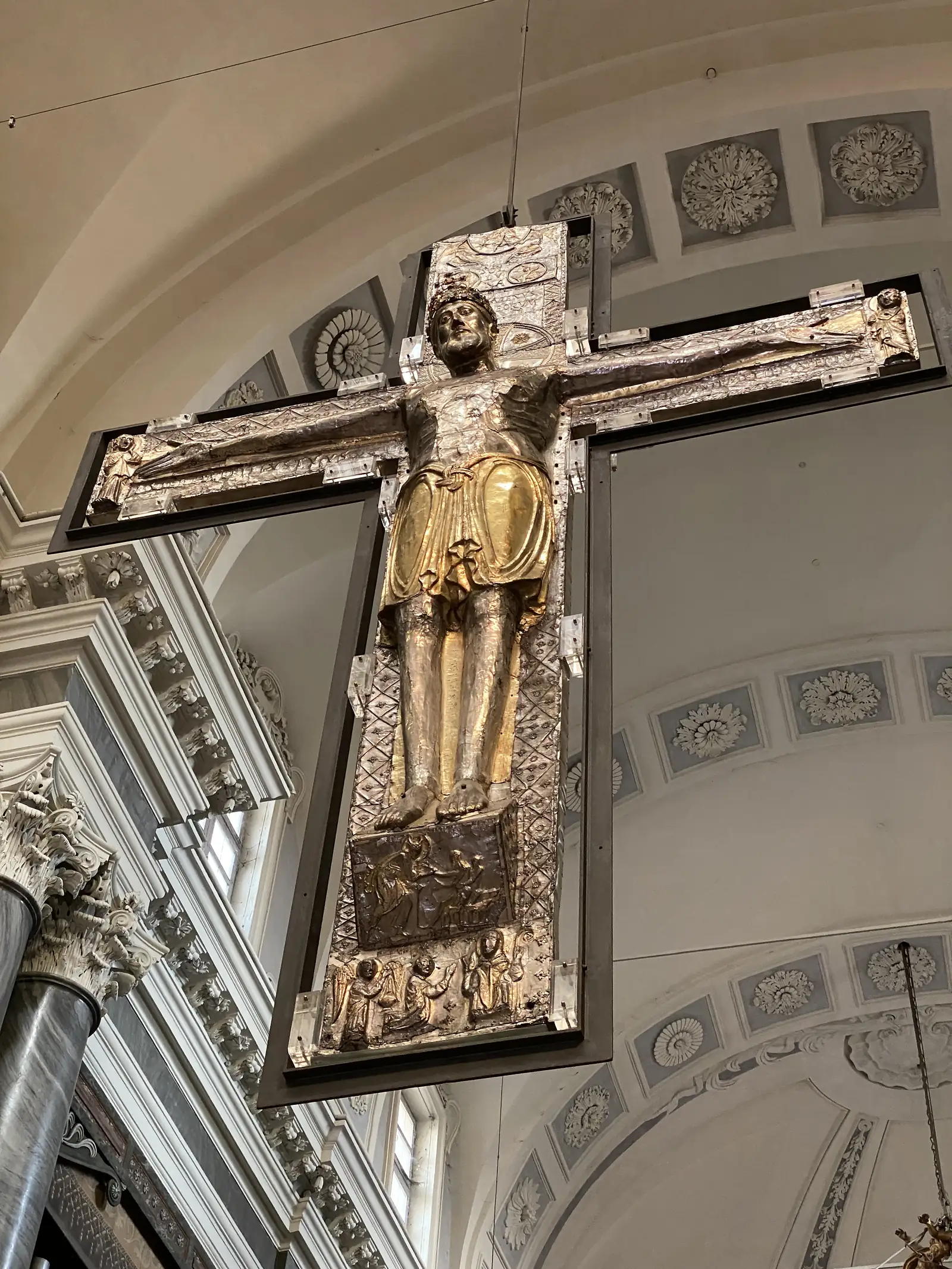 A large gilded and silvered crucifix suspended beneath a coffered dome, its figure of Christ picked out in gold against the ornate plasterwork of the church ceiling.