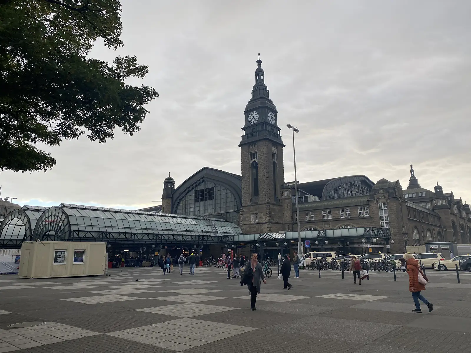 A grand European railway station with a tall clock tower rises behind a paved square where pedestrians cross under an overcast sky.