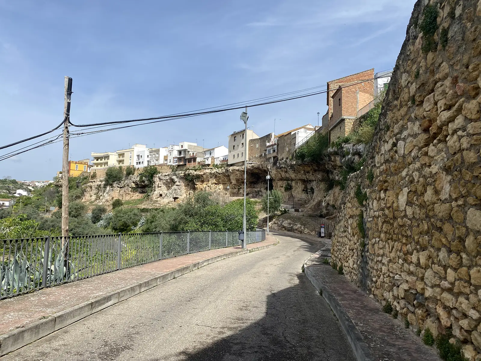 A road curving beneath a massive rough stone retaining wall, with houses perched along the edge of the limestone cliff above.