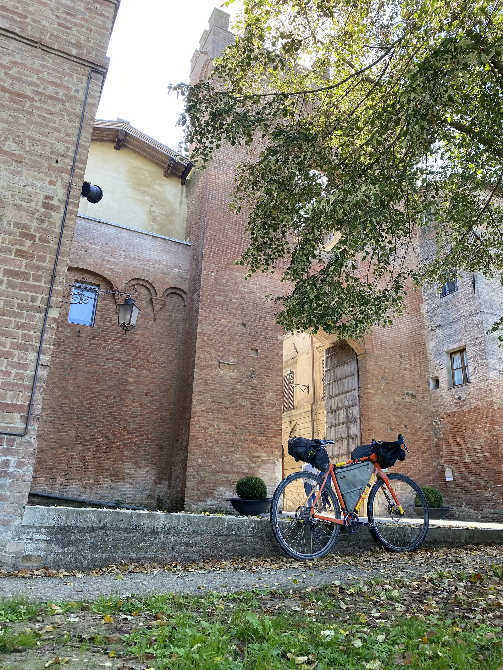A loaded gravel bike propped against a wall in a brick-paved lane between old town buildings, with a wrought-iron lamp bracket nearby.