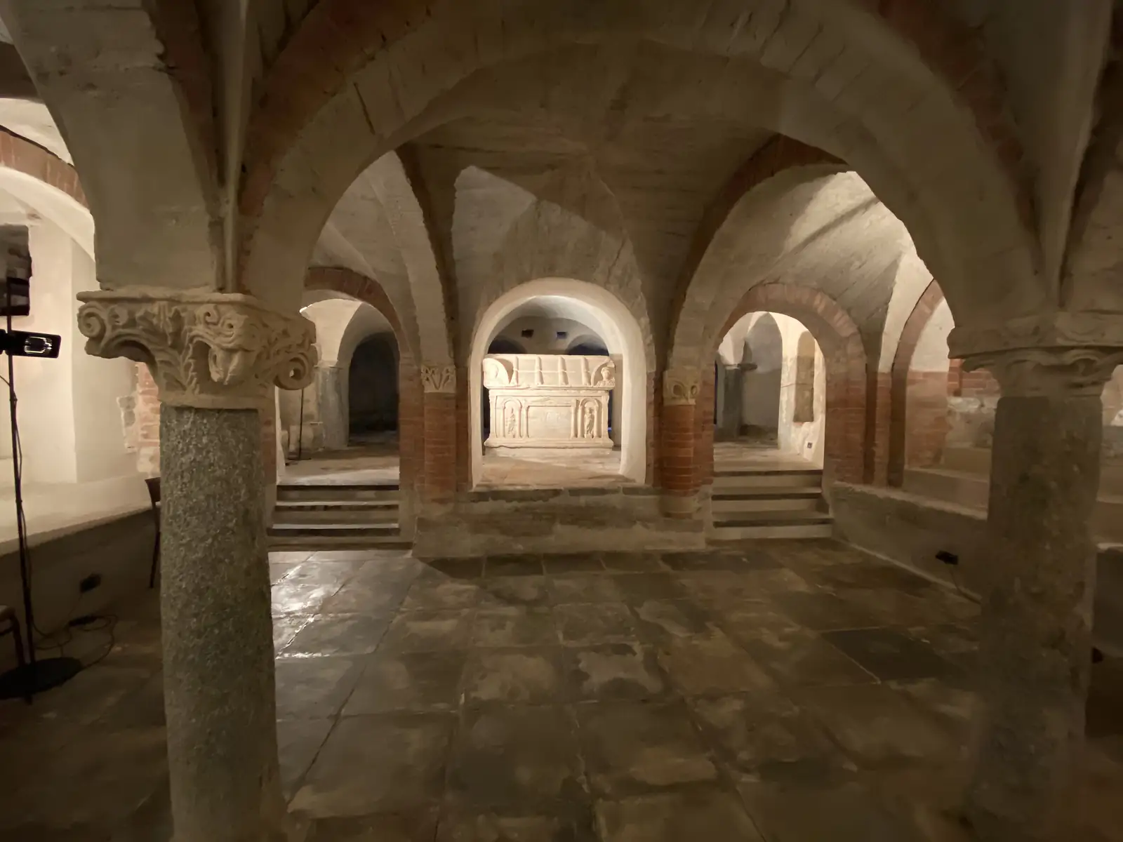 A vaulted Romanesque crypt with squat stone columns and carved capitals, its arches framing a raised stone sarcophagus lit at the centre of the space.