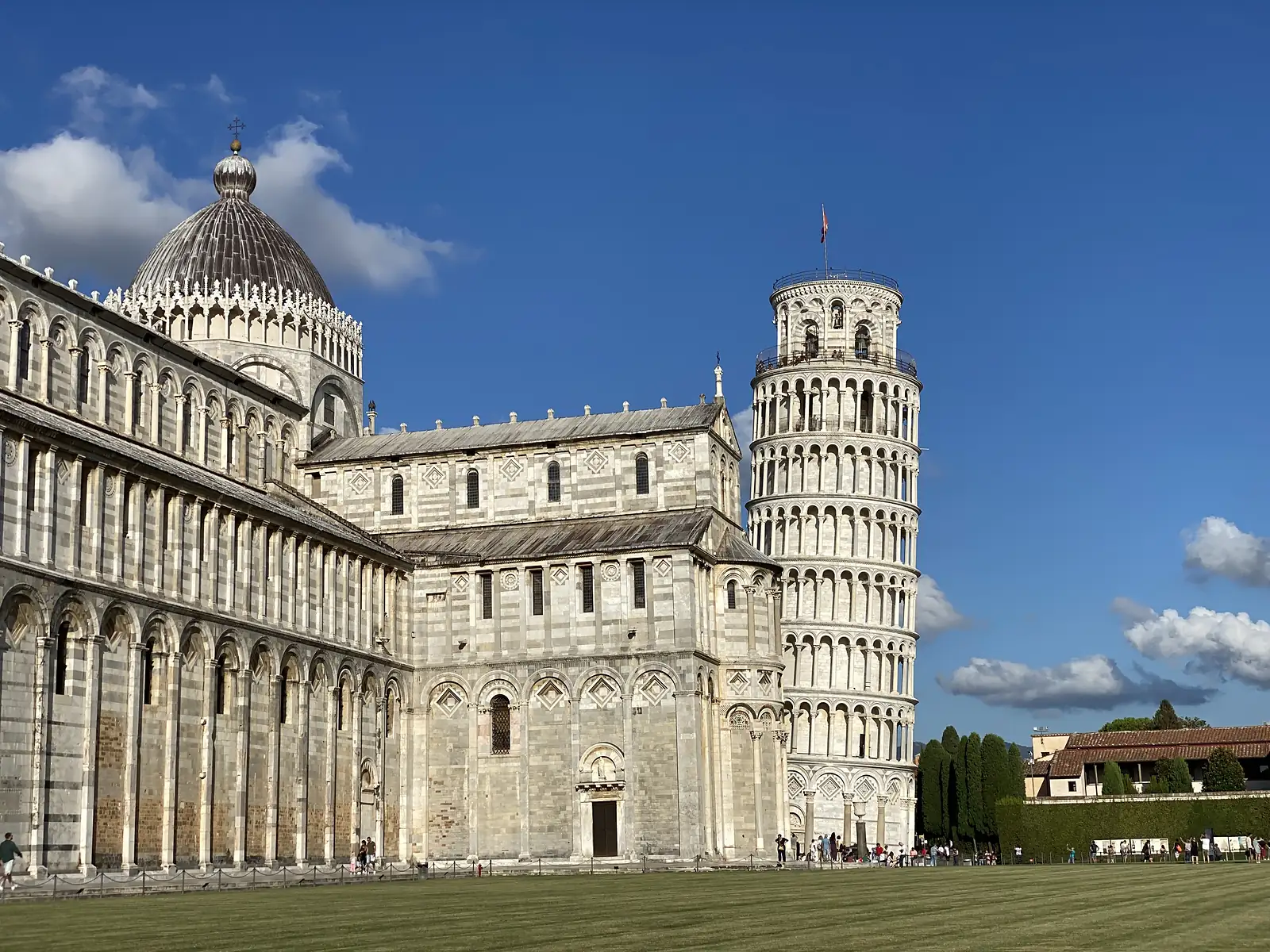 The striped dome of Pisa Cathedral and the tilted Leaning Tower rise side by side against a clear blue sky, a flag flying from the tower's summit.