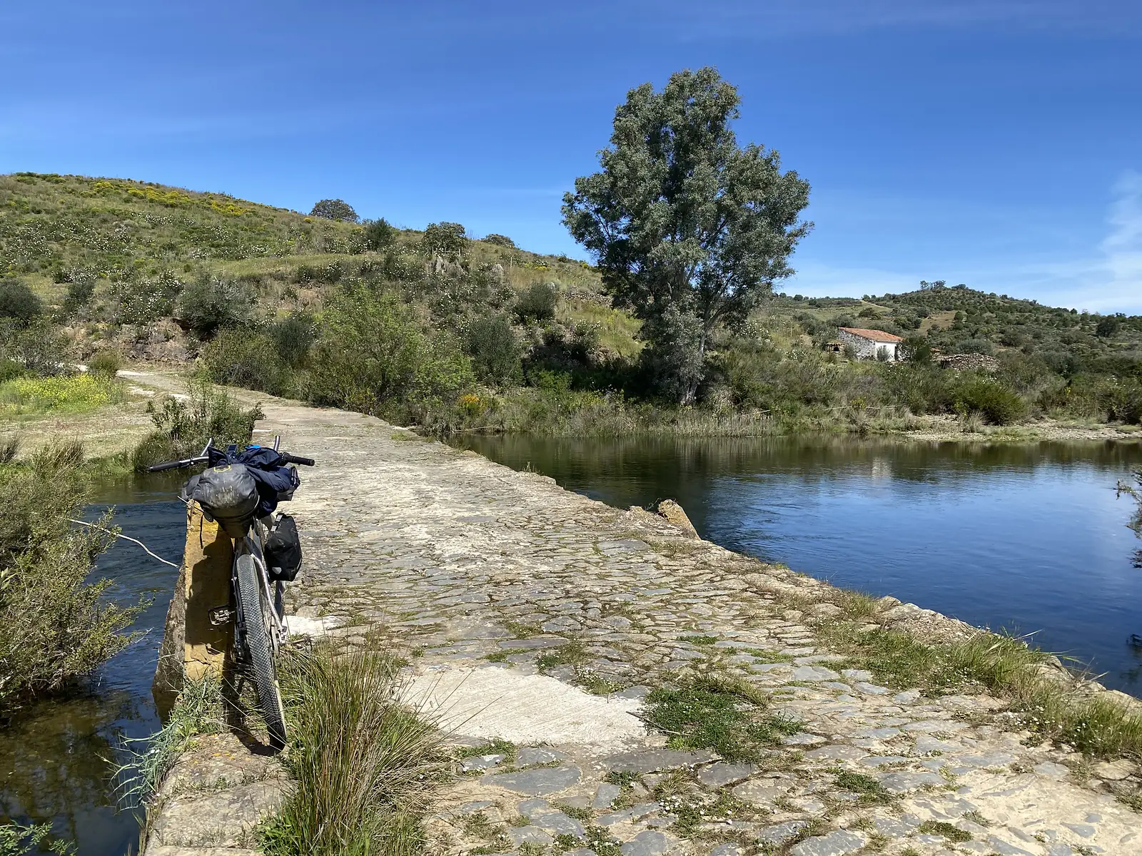 A loaded bikepacking bike propped against the parapet of a low cobbled bridge across a calm river, with scrubby hills and a white farmhouse on the far bank.