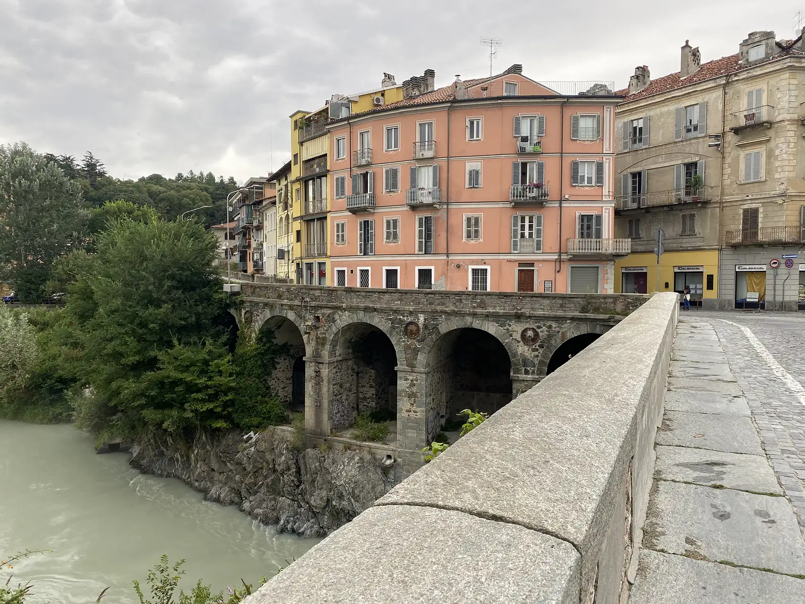 A stone arched bridge crossing a river gorge, with a pink-painted apartment building and older townhouses lining the far bank of the town.