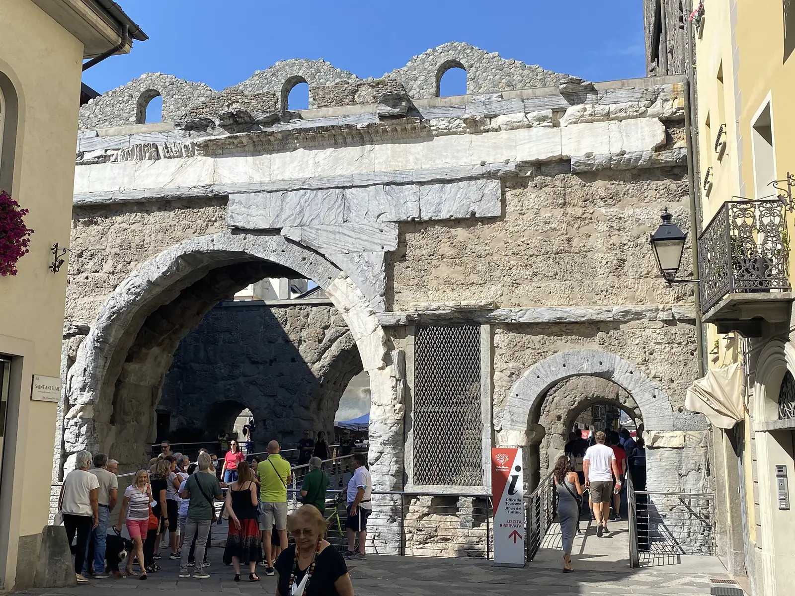 Pedestrians pass beneath twin stone archways of an ancient town gate, its weathered walls topped with a jagged ruined parapet against a partly cloudy sky.