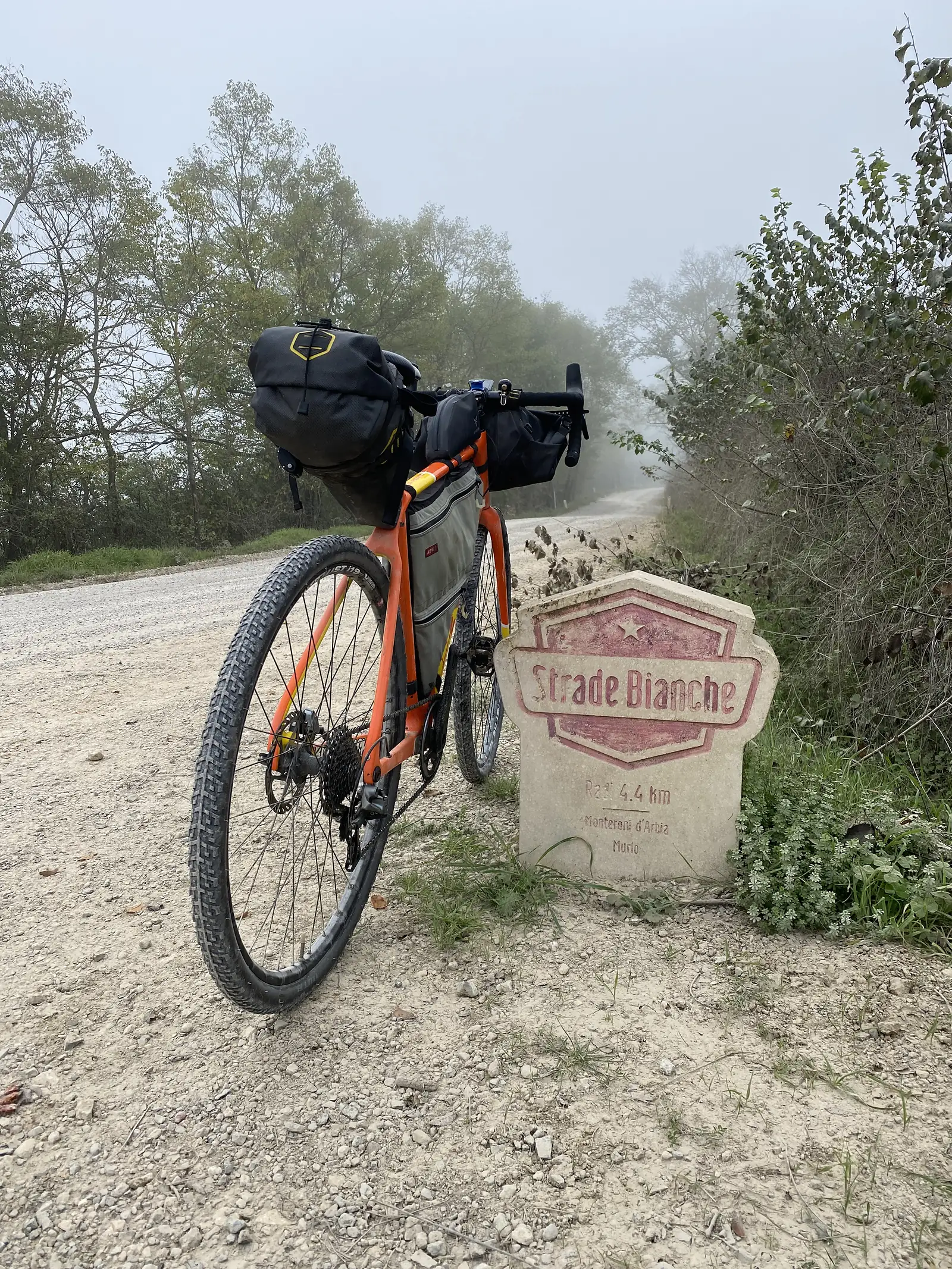A loaded orange gravel bike leaning against a stone marker inscribed "Strade Bianche" beside a white gravel road in fog.