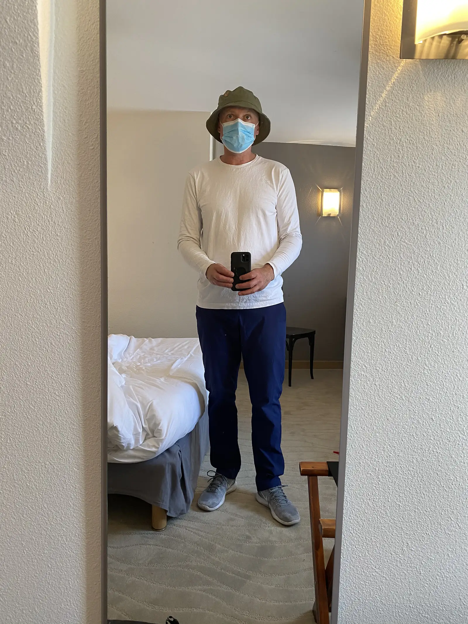 A cyclist wearing a bucket hat and surgical face mask takes a mirror selfie in a plain hotel room, an unmade bed behind him.