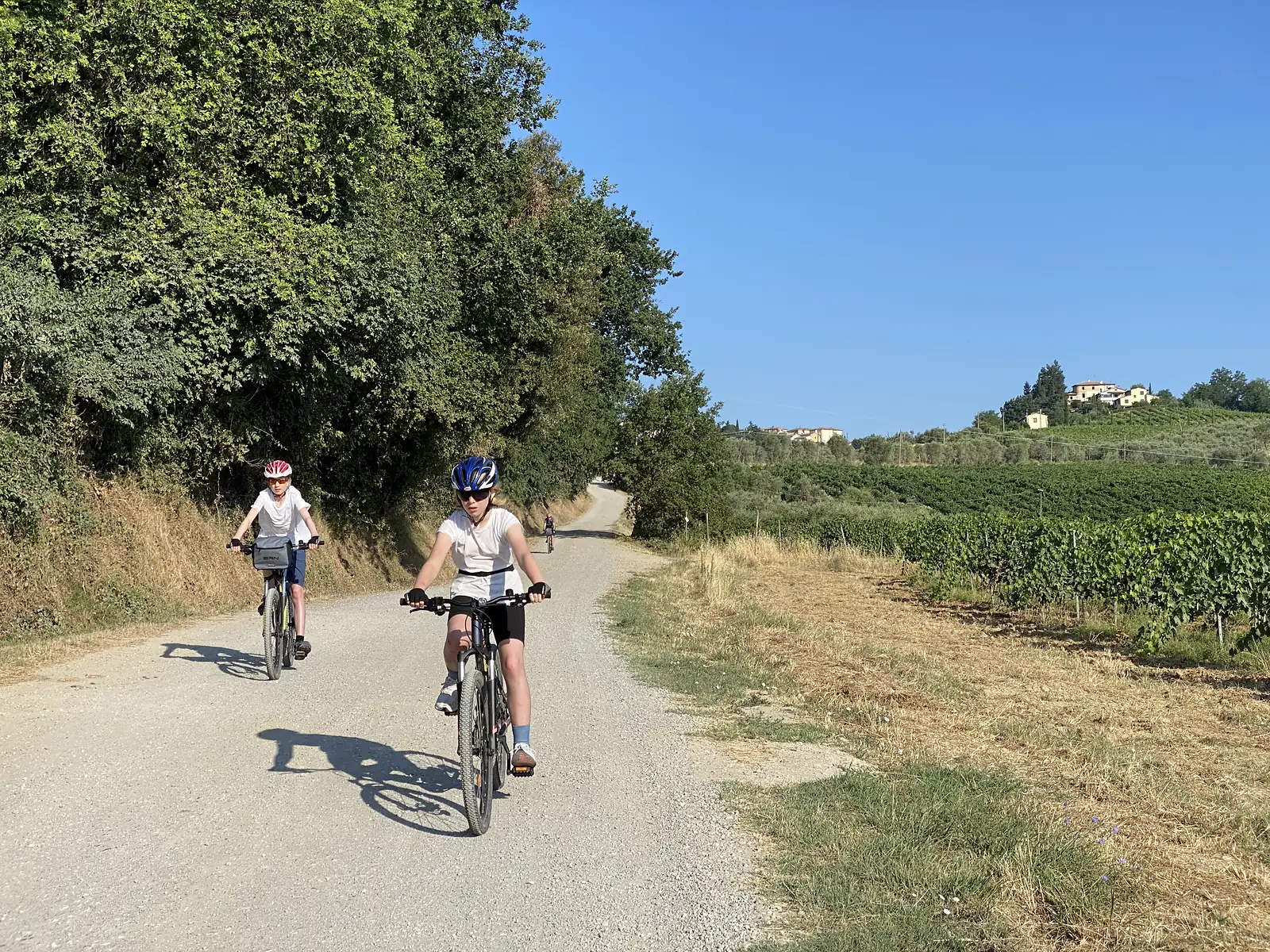 Two young cyclists in helmets ride a gravel lane hedged with trees, with vineyards and a hilltop hamlet visible ahead.