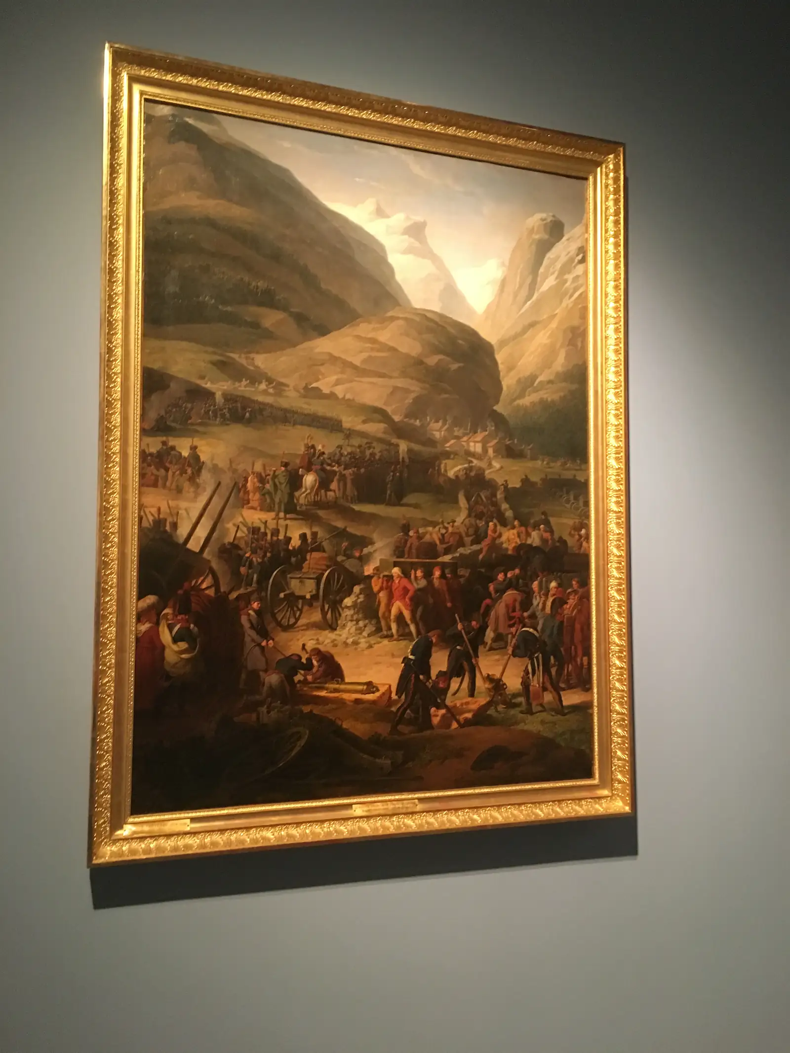 A gilt-framed painting of an army crossing a mountain pass, soldiers, cannon and pack horses winding along a rocky track beneath towering peaks.