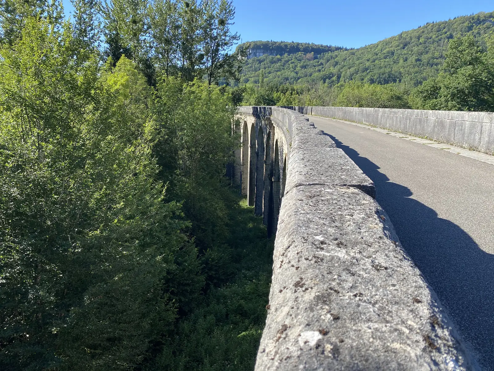 A long stone viaduct with a series of arches carries a road above a wooded valley, forested hills rising beyond.