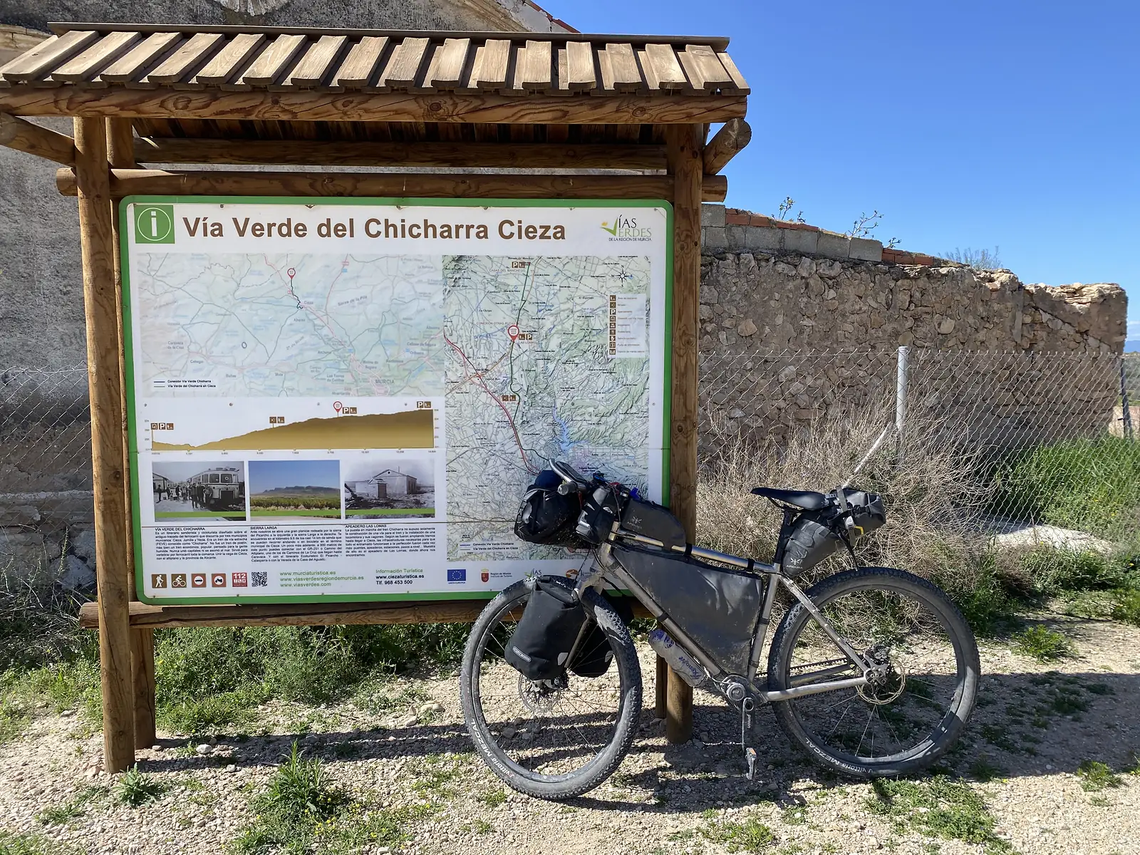 A mud-spattered loaded bikepacking bike leaning against a wooden information board headed "Via Verde del Chicharra Cieza", with route maps and a height profile.