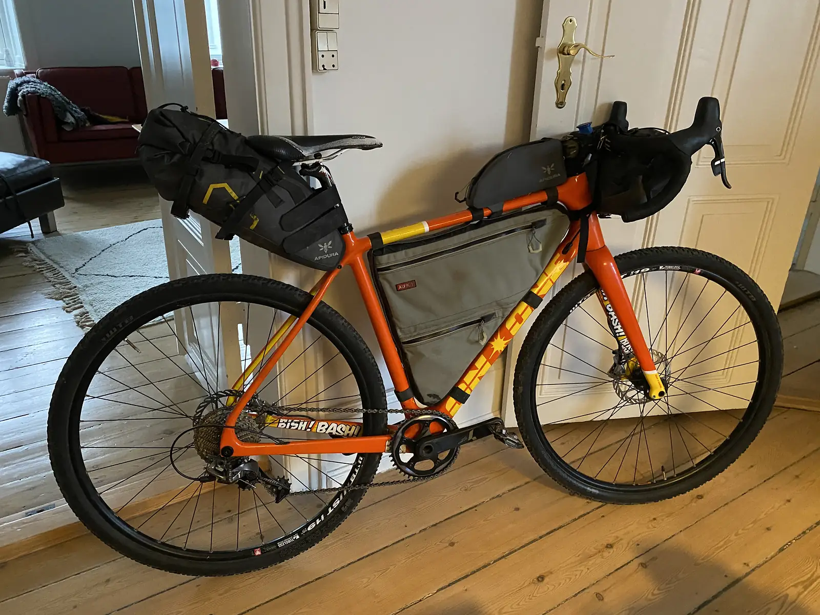 A loaded orange gravel bike with bikepacking bags leans against a door indoors, its frame lettered 'BISH! BASH!'