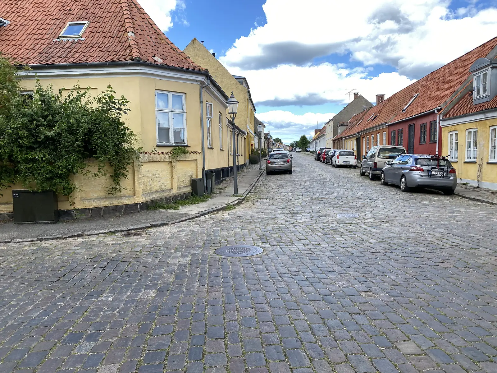 A wide cobbled street corner of yellow, red and ochre rendered houses with red-tiled roofs and cars parked along it.