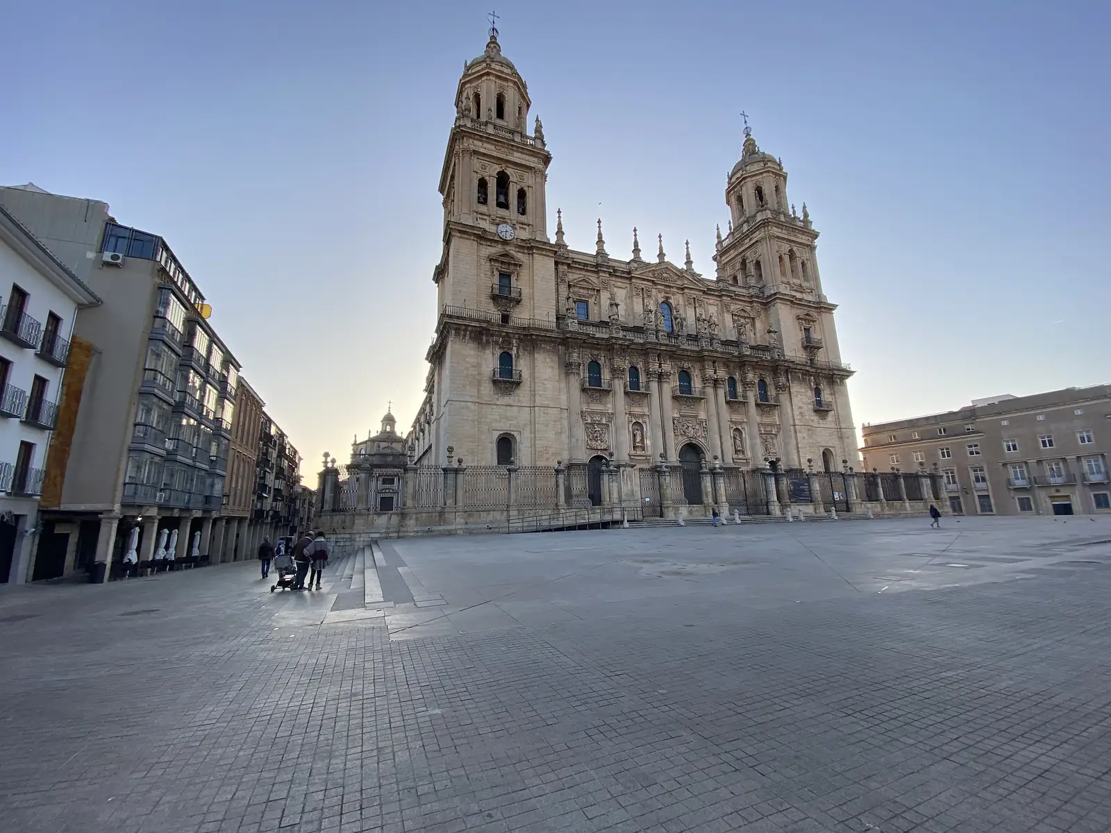 A vast baroque cathedral front with twin bell towers and ranks of statues, seen across an empty paved square at first light.