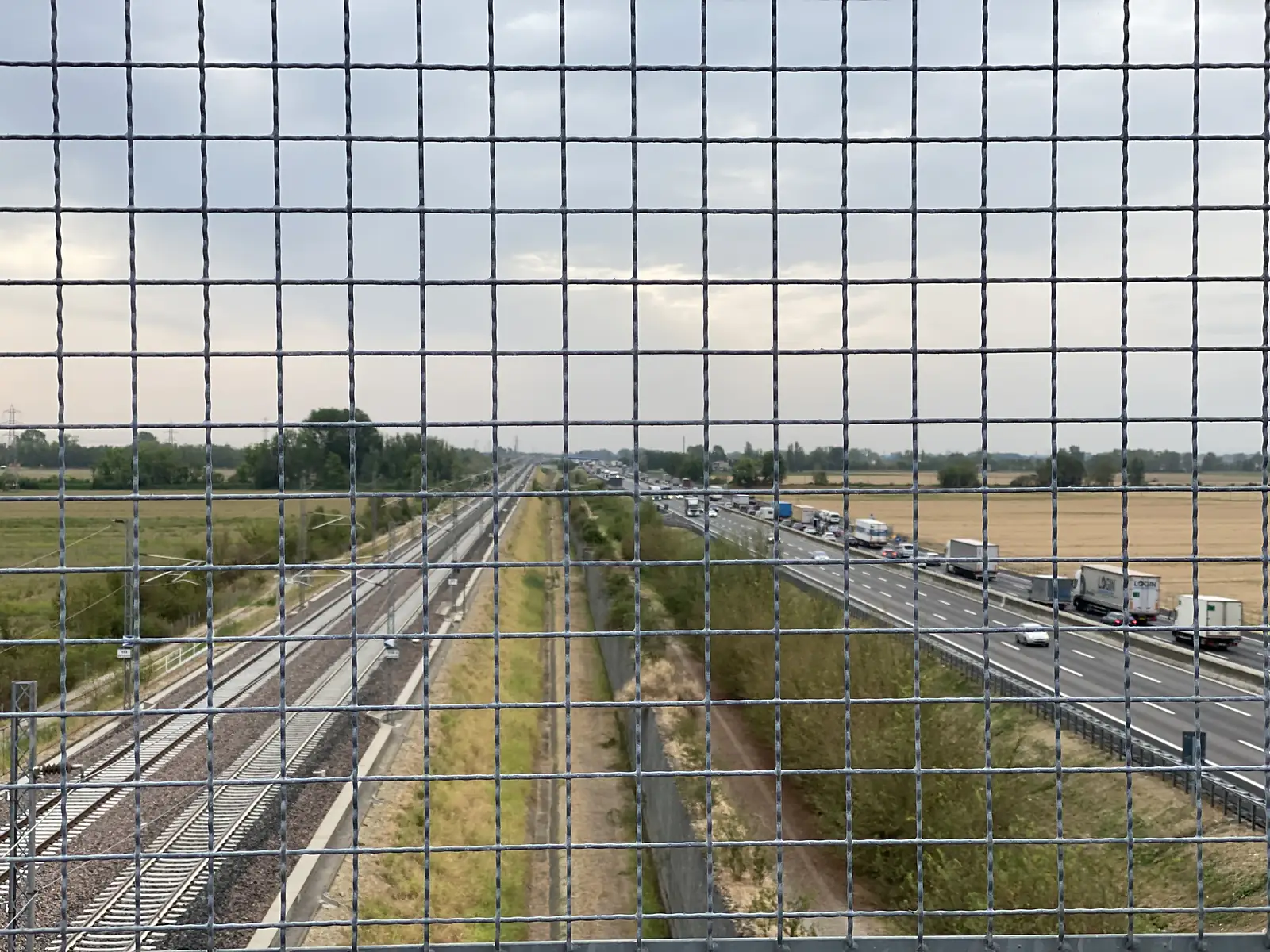 A view through a level-crossing style mesh fence down onto a railway line running beside a busy motorway with queued lorries and cars.