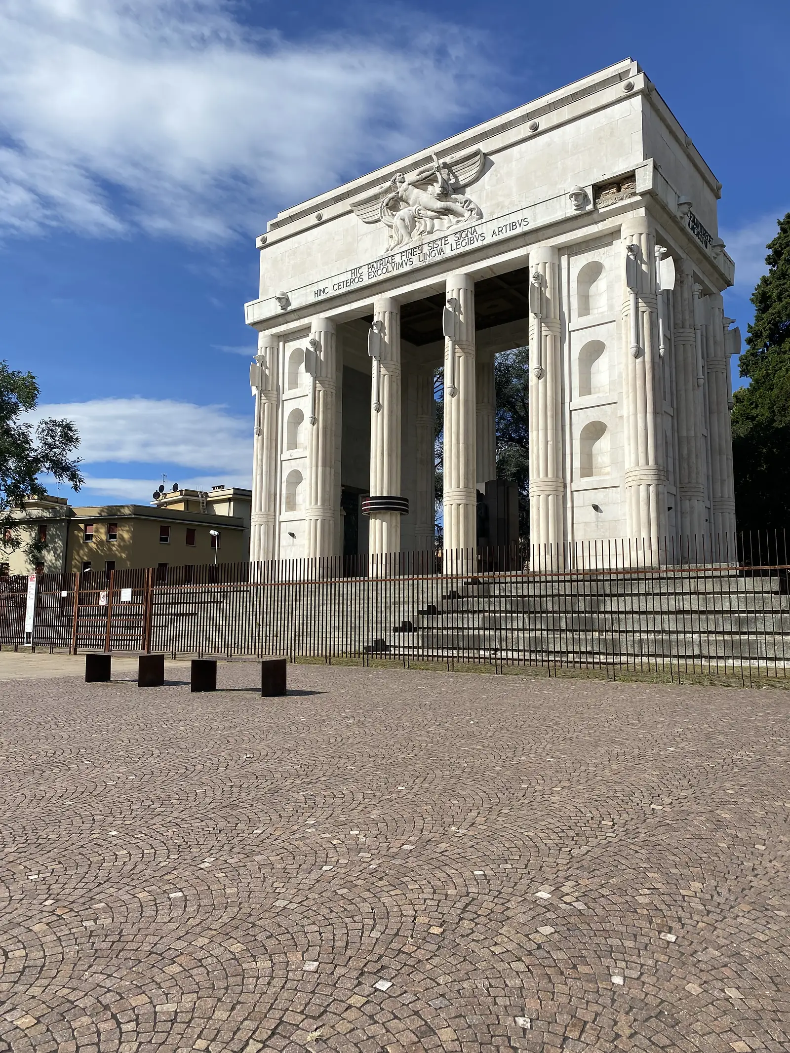 A monumental white stone arch with fluted columns, a winged relief and a Latin inscription stands behind railings on a cobbled square.