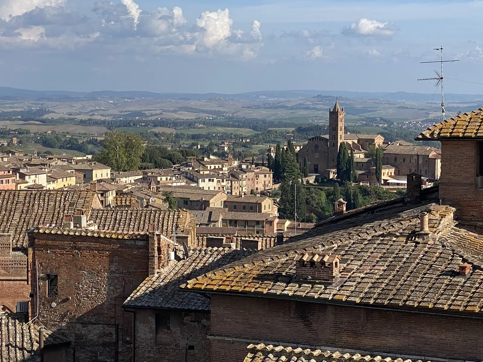 Terracotta rooftops and a church tower of a Tuscan hill town, with the surrounding countryside stretching to distant hills beneath a partly cloudy sky.