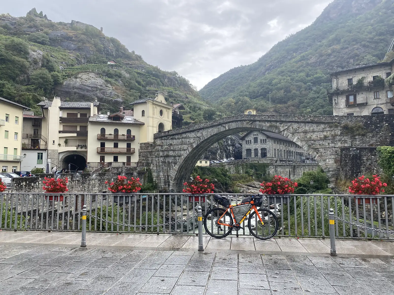 A loaded orange gravel bike leaning against a riverside railing hung with red flower baskets, with a tall stone arch bridge and an old stone building beyond.
