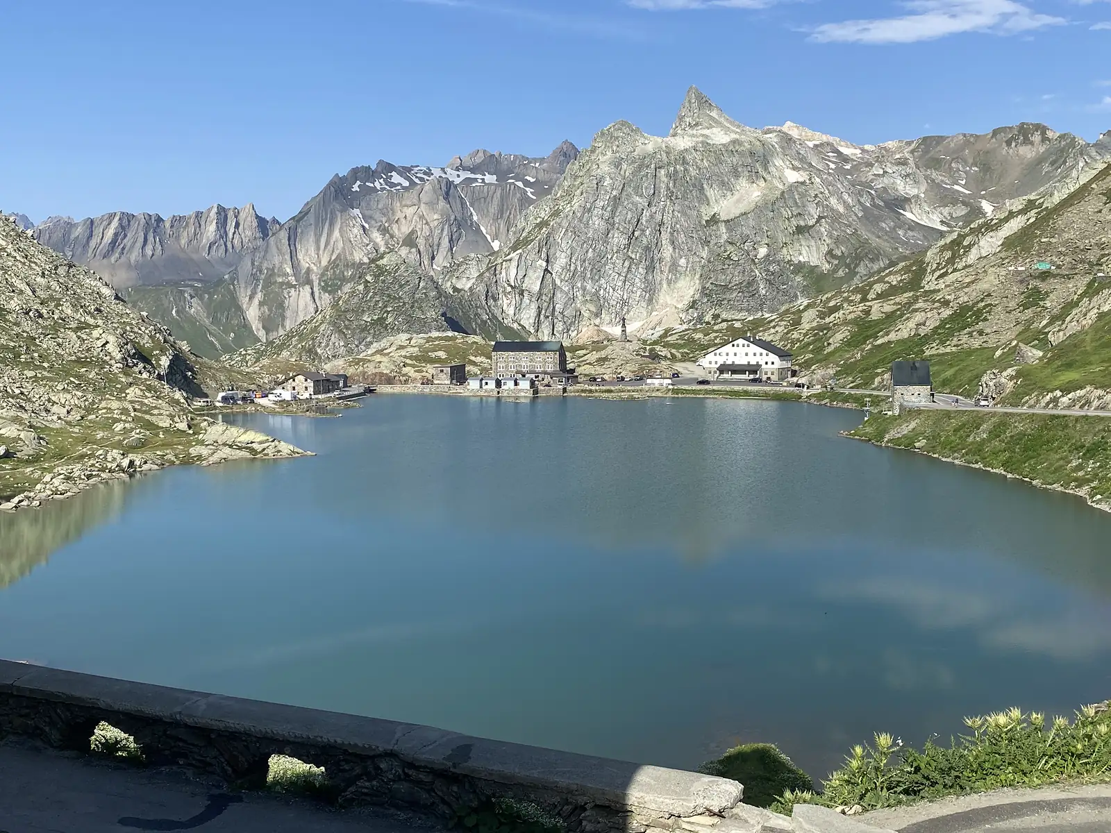 The Grand-St-Bernard lake sits beneath a jagged granite peak, with the hospice and monastery buildings ranged along its far shore.