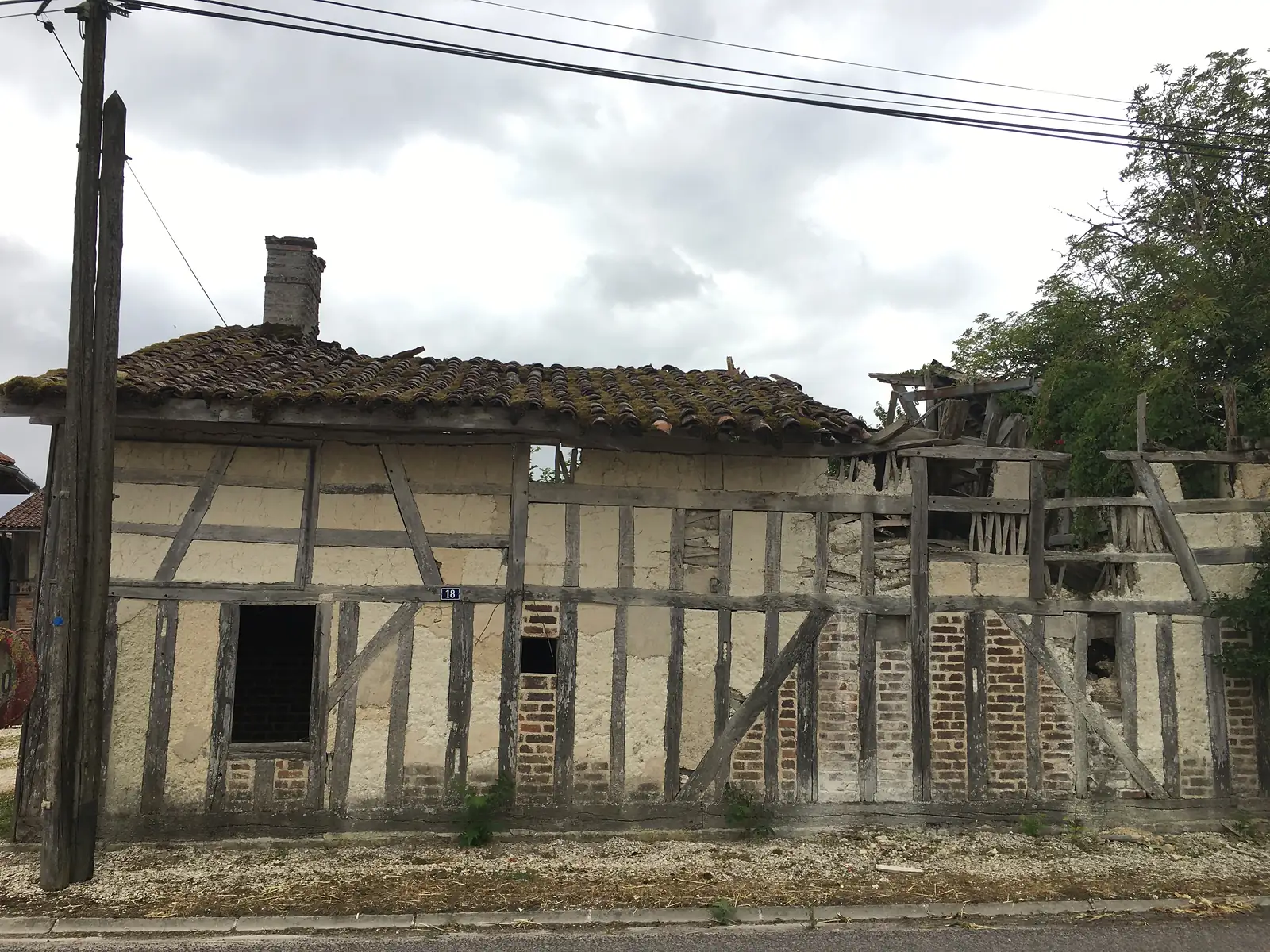 A part-collapsed timber-framed cottage numbered 18 stands roofless at one end, its render crumbling from the exposed frame, on a village street.
