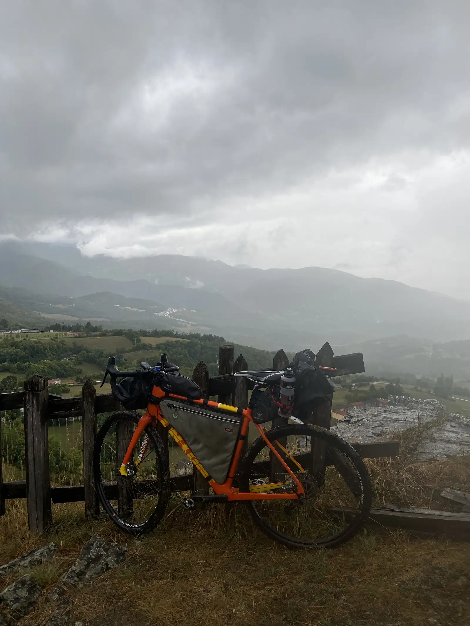 A loaded orange gravel bike with a frame bag and bar bag leans against a wooden fence, overlooking a hillside village under heavy cloud.