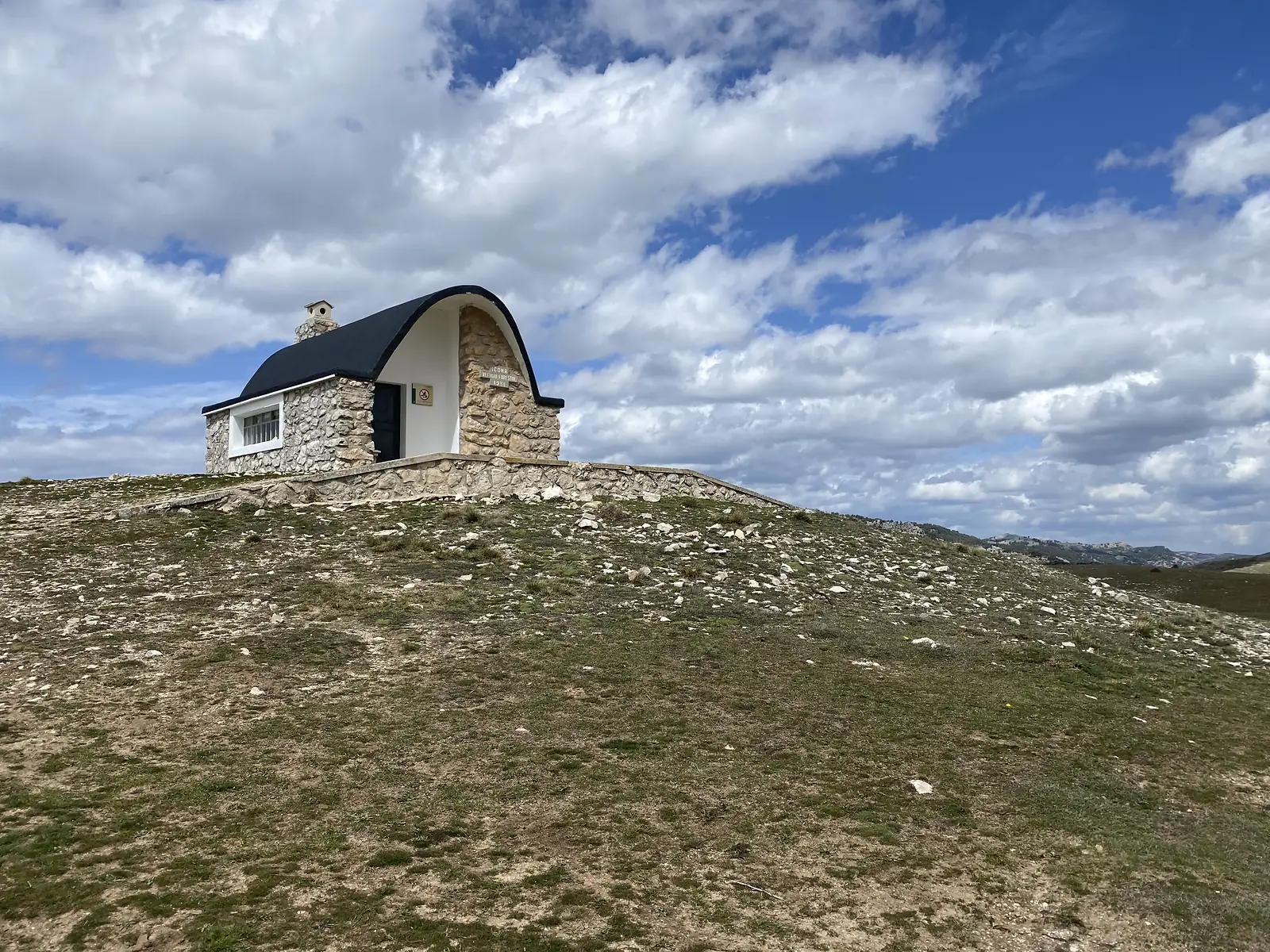 A stone mountain refuge with a curved slate roof standing alone on a bare grassy rise, a plaque above its door.