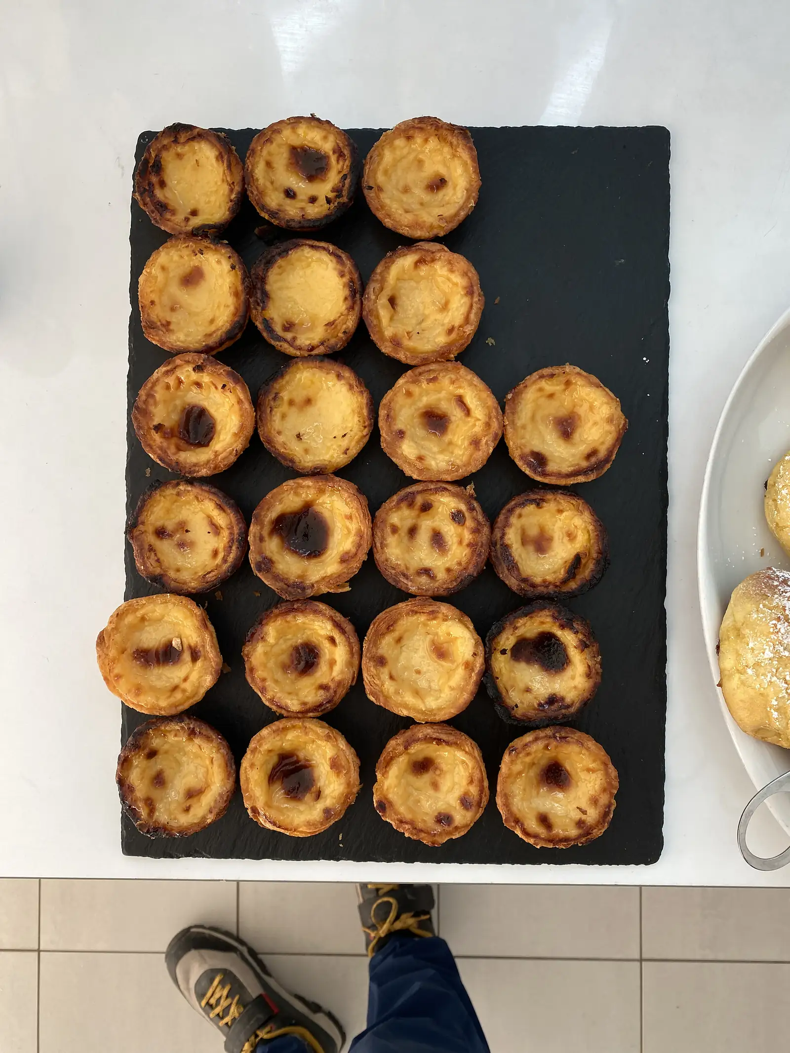 Slate board holding twenty-two custard tarts with scorched, blistered tops, a plate of sugar-dusted pastries alongside, photographed from above with the toes of a pair of cycling shoes in the frame.