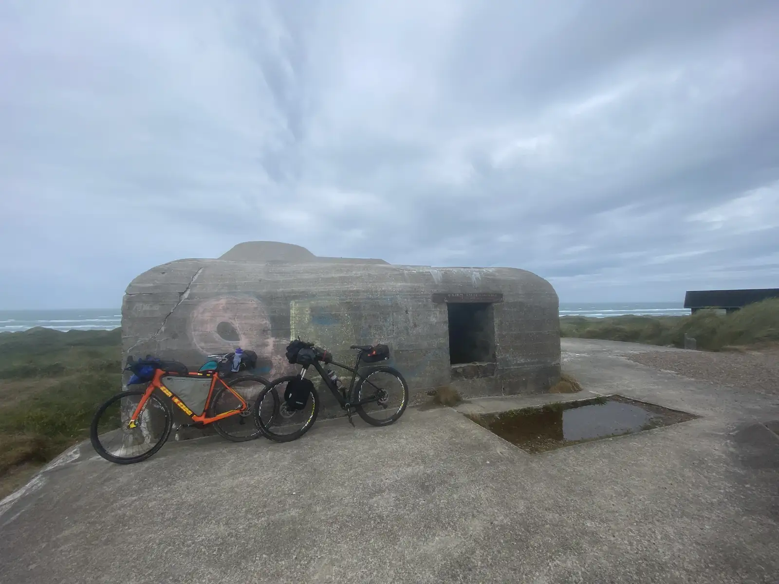 Two loaded bikes, one orange and one black, leaning against a graffiti-marked concrete bunker on a dune clifftop above a grey sea.
