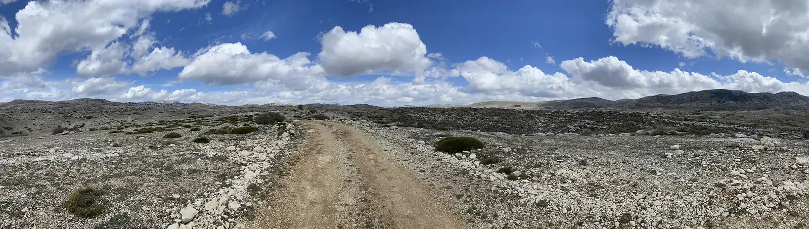 An ultra-wide view of a stony track crossing a bare limestone plateau beneath towering white cumulus.