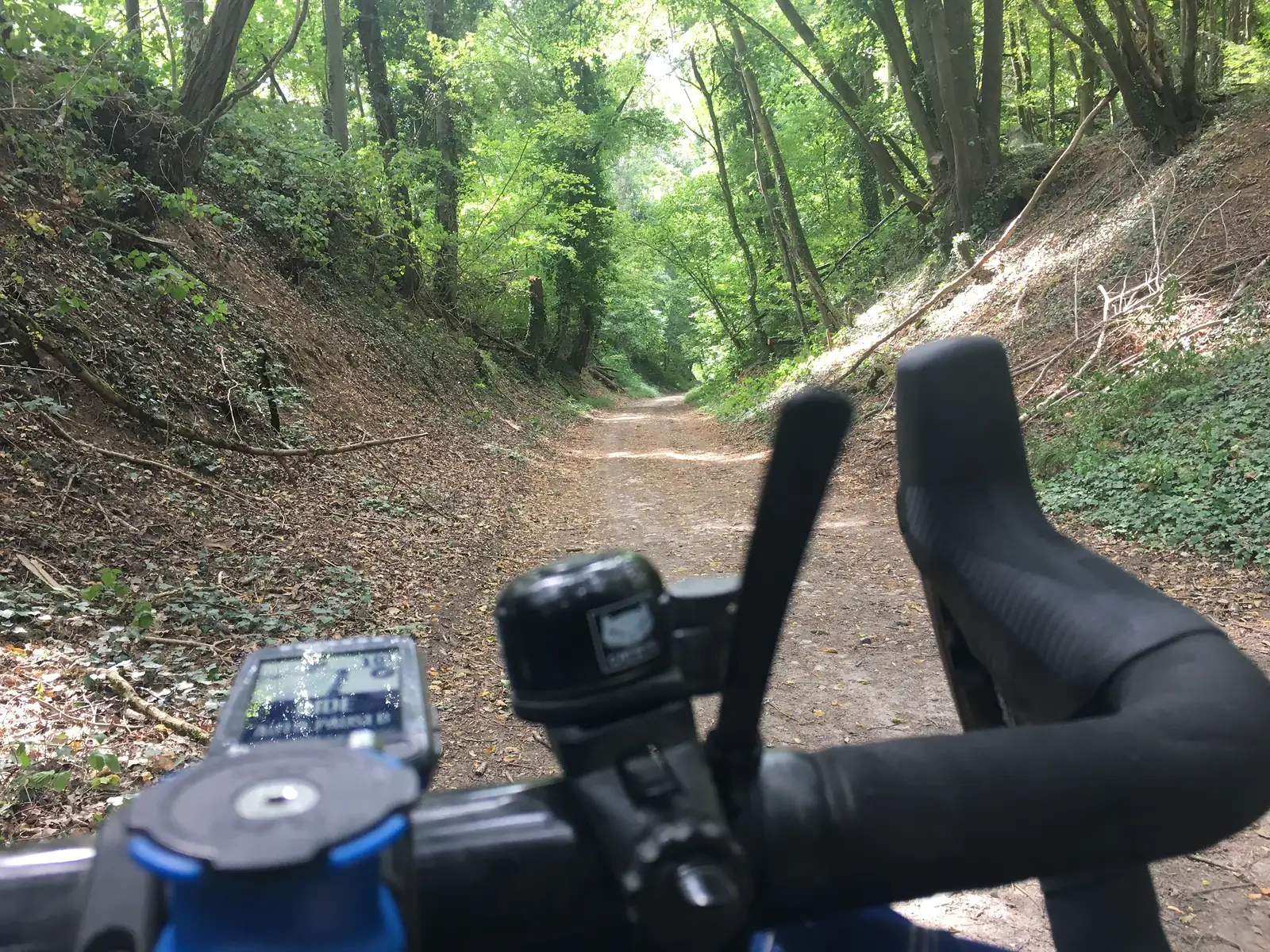 A cyclist's-eye view along a sunken, leaf-strewn forest track between earth banks, handlebars and a cycling computer visible in the foreground.
