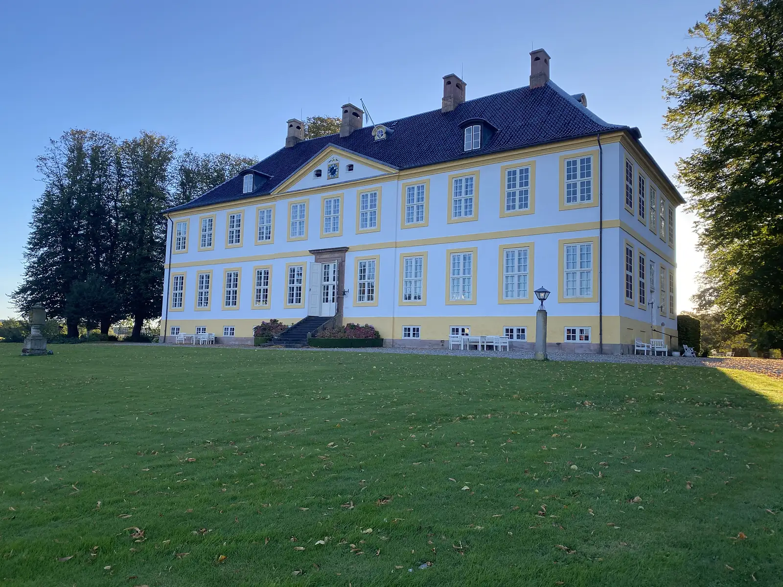 A grand white and yellow baroque manor with a pedimented centre, clock and tall chimneys standing above a wide lawn.