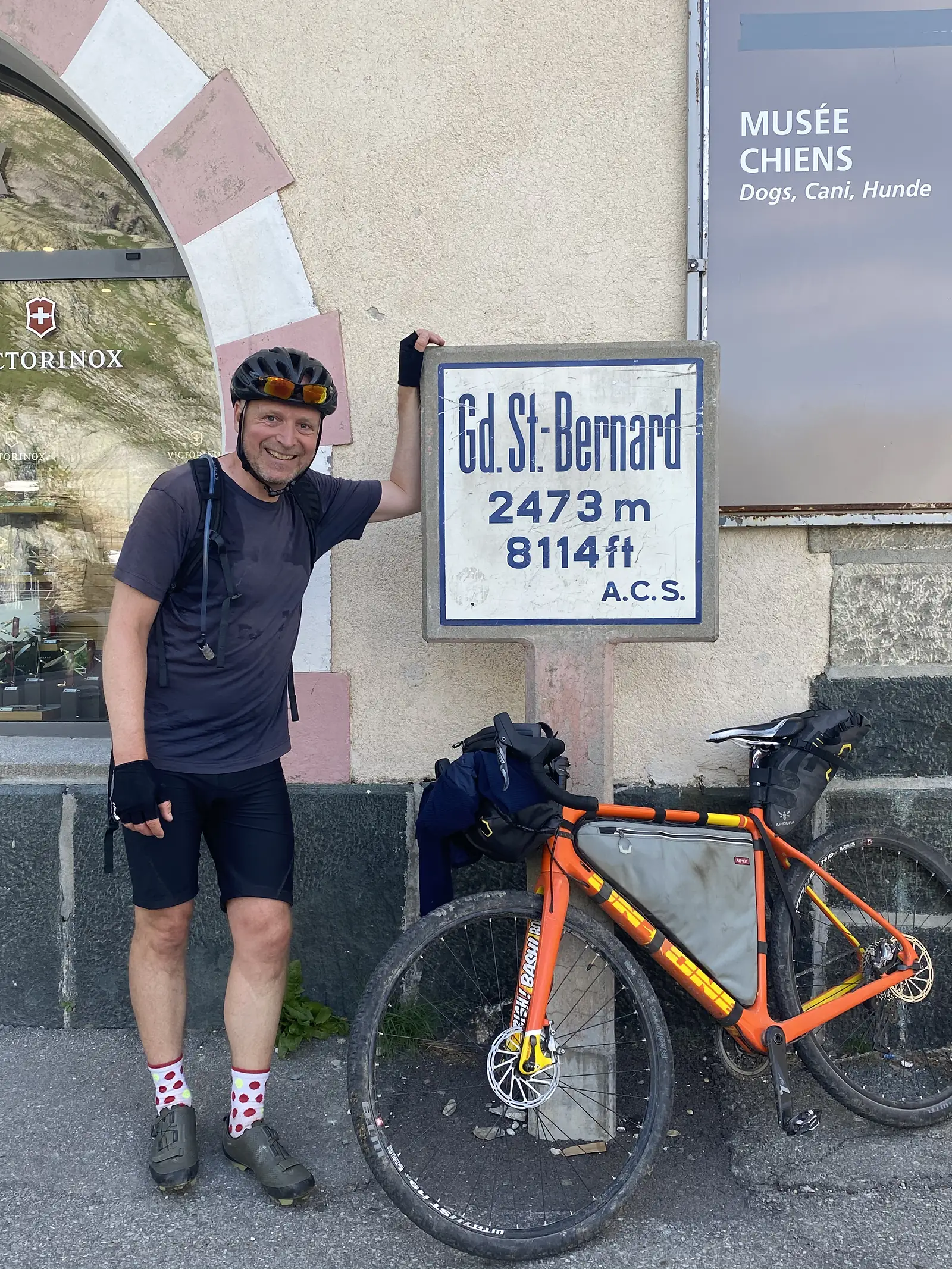 A cyclist stands beside his orange loaded touring bike with one hand on a stone marker reading 'Gd. St-Bernard 2473m 8114ft', outside a shopfront at the pass.
