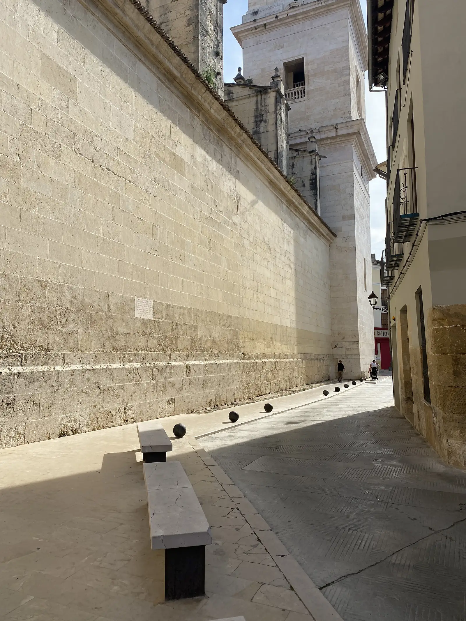A narrow lane in deep shade beside a church's long ashlar flank, with stone benches and a bell tower rising at the end.