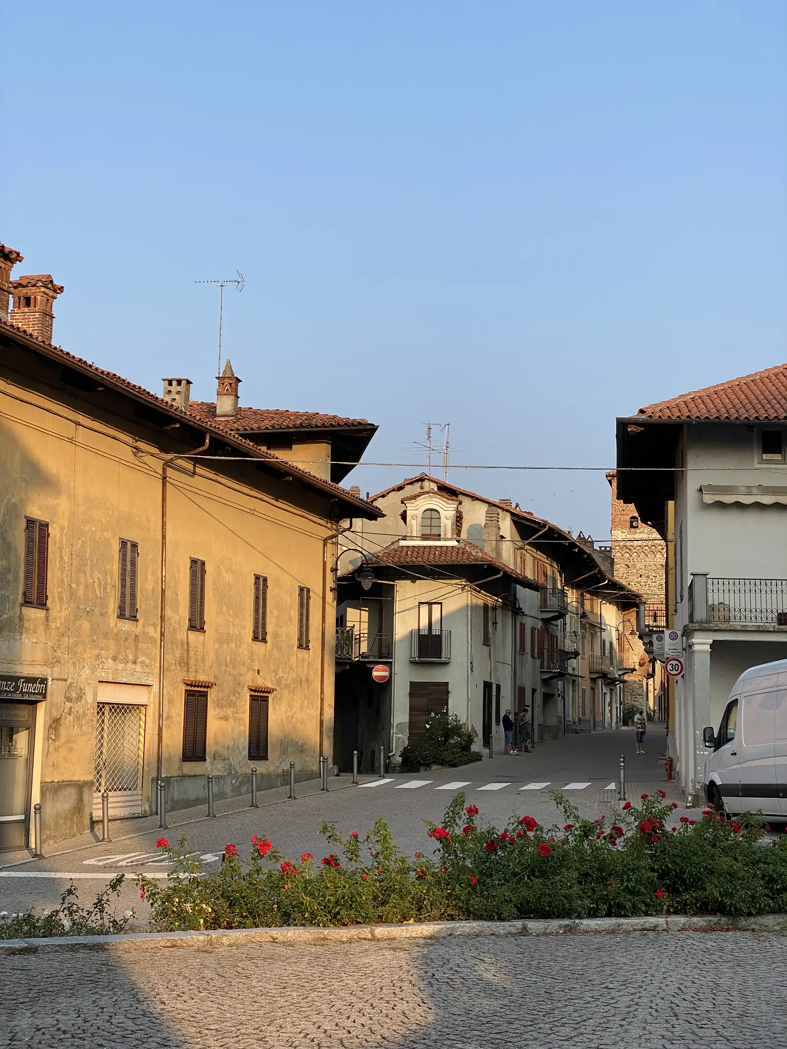 A quiet cobbled street bathed in evening light, its ochre buildings edged by a low wall thick with red roses.