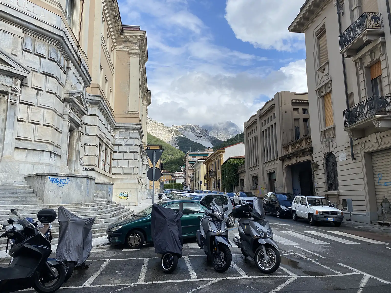 A street of grand stone-faced townhouses opens onto scarred white marble quarries on the mountainside beyond, with mopeds and cars parked along the kerb.