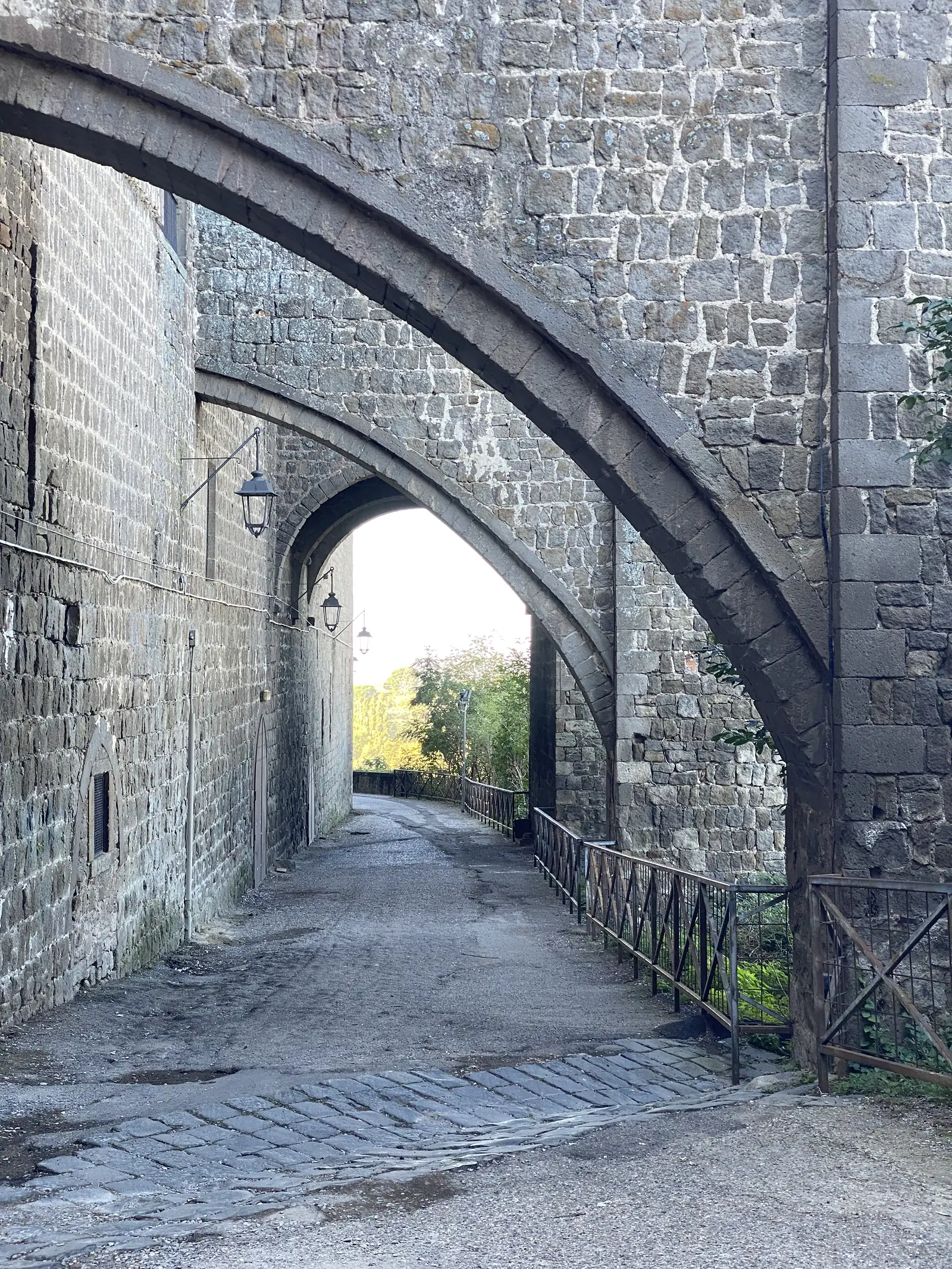 A stone-vaulted passageway beneath arched buttresses, cobbled underfoot with wall-mounted lanterns leading towards daylight at the far end.