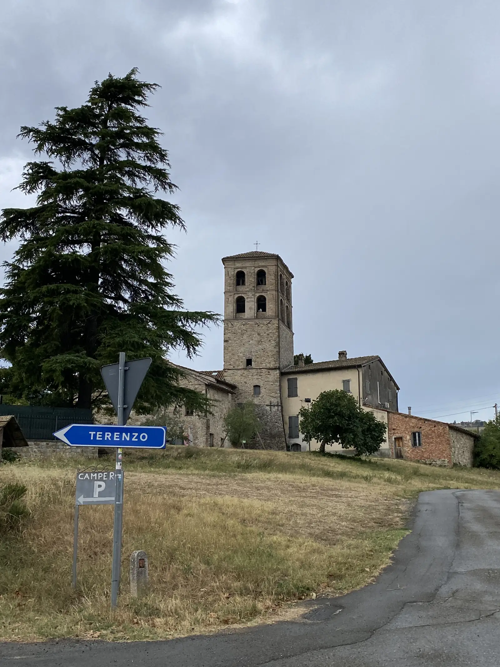 A blue road sign reading 'Terenzo' with a camper-parking symbol stands before a stone bell tower and cluster of village houses on a wooded hillside.