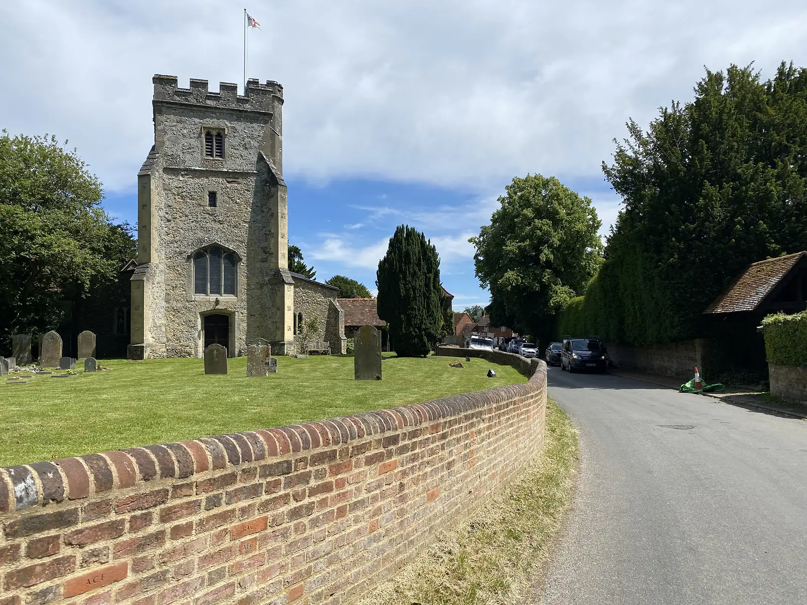 A flint and stone church with a battlemented tower flying the St George's flag, above a brick churchyard wall and headstones.