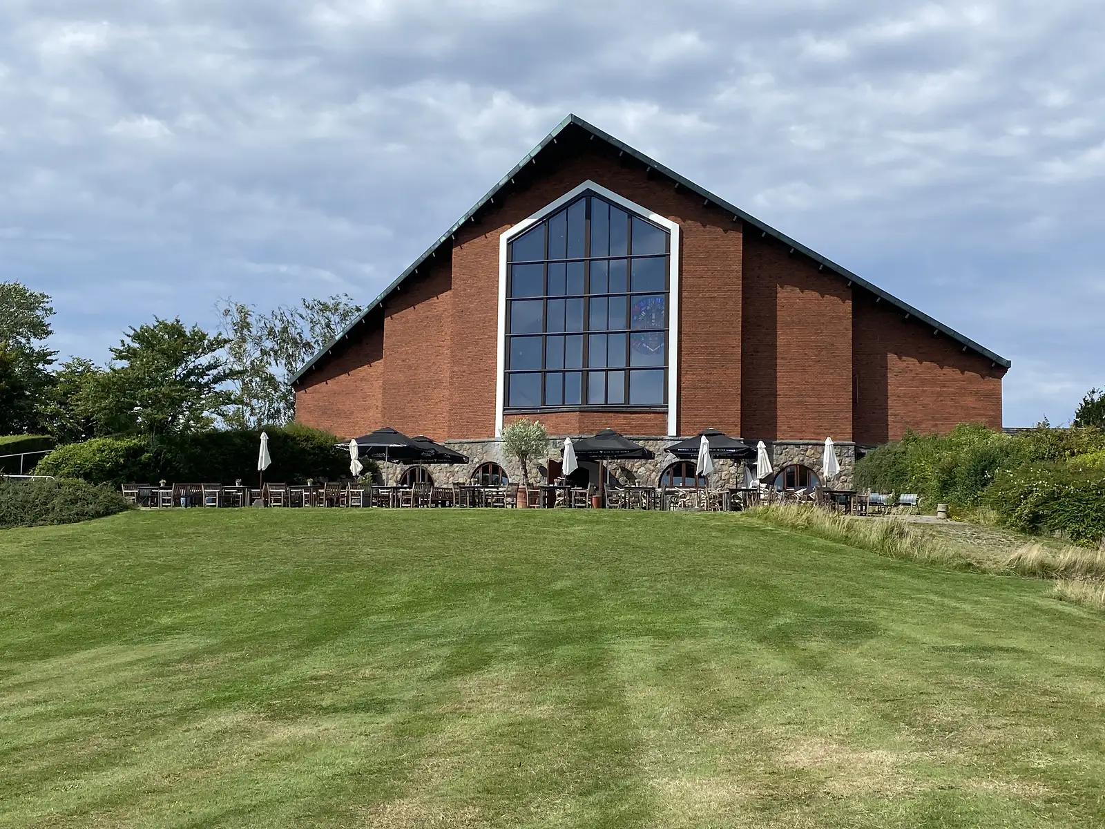 A large red-brick building with a steep gable and tall arched window above a mown lawn, parasols and tables outside.